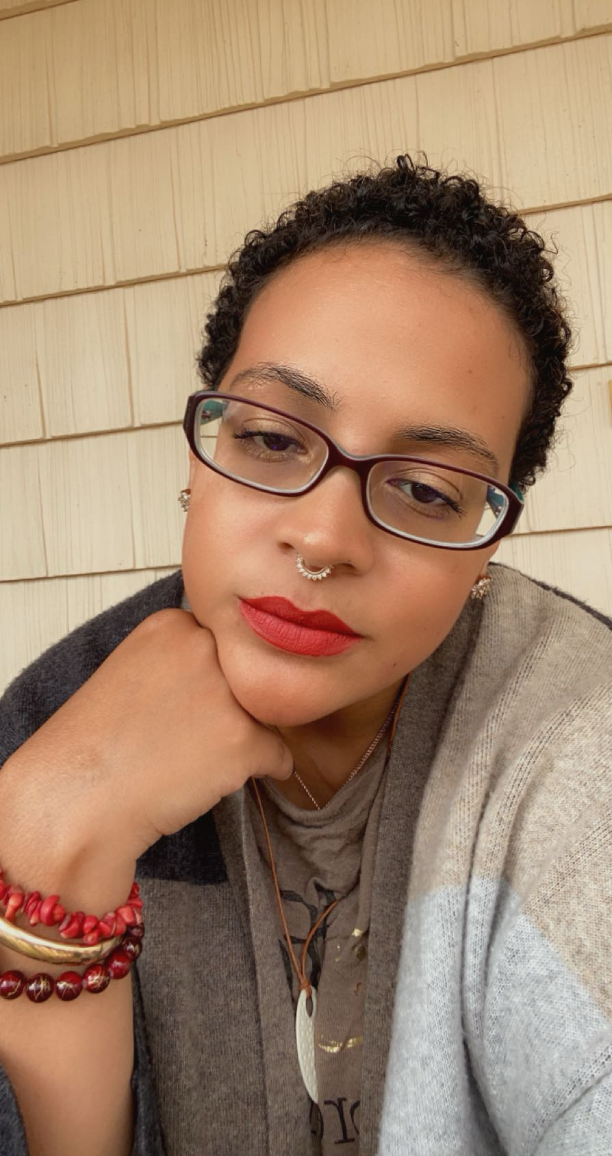 Woman with short curly hair, glasses, red lipstick, and red bracelets resting her chin on her hand, against a wooden wall.