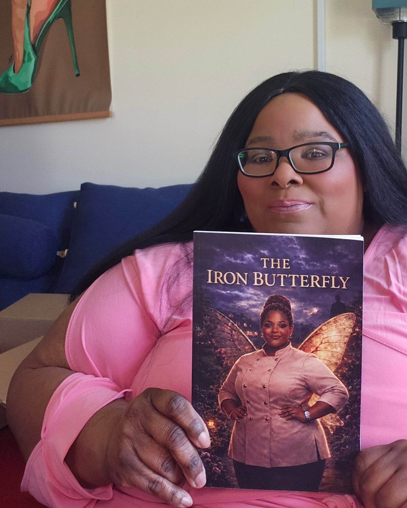Woman with glasses in pink shirt holding a book titled 'The Iron Butterfly' with a fairy on the cover.