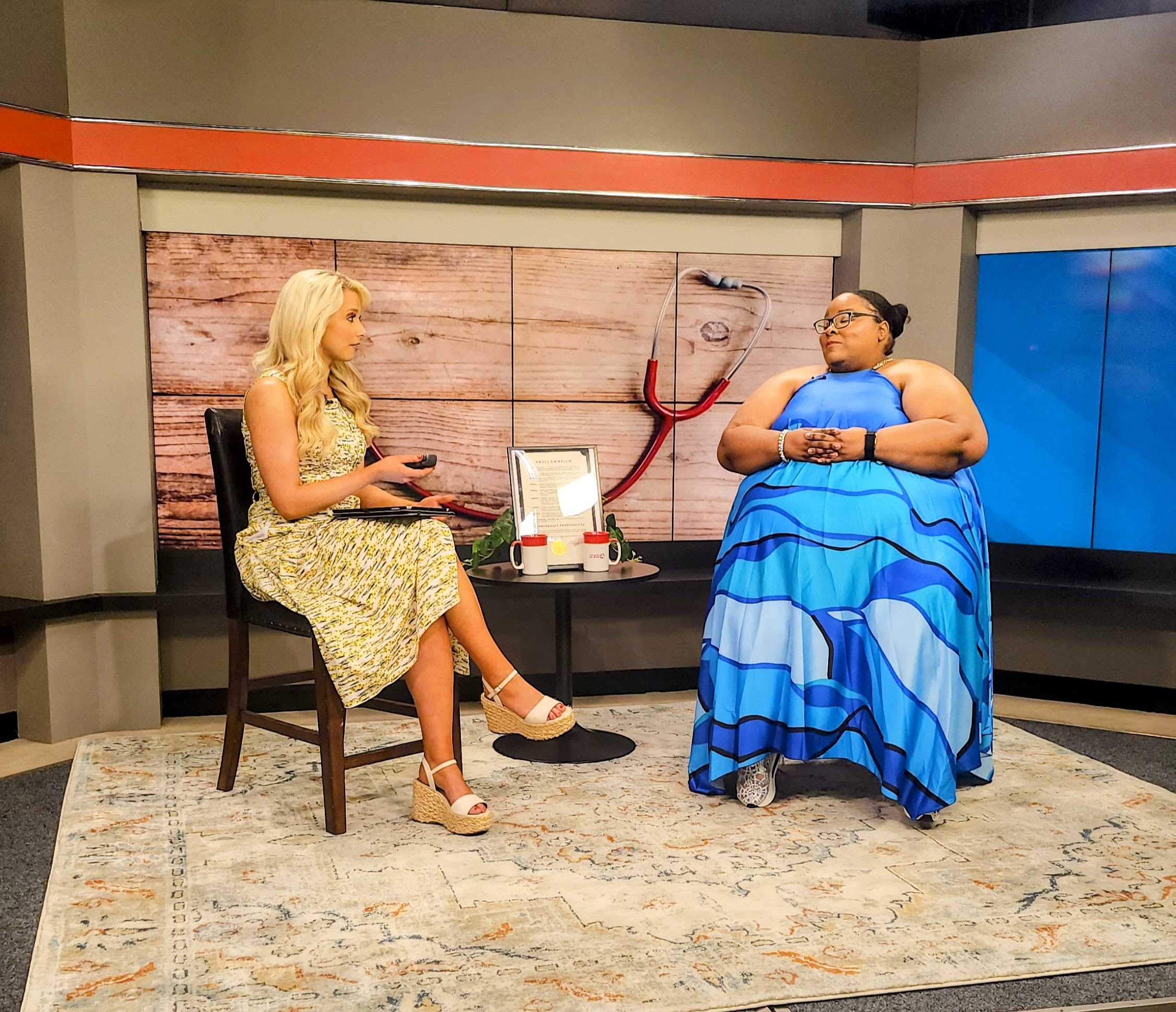 Two women sit and talk in a studio with a wooden backdrop and a blue screen, one in a floral dress, the other in a blue dress.