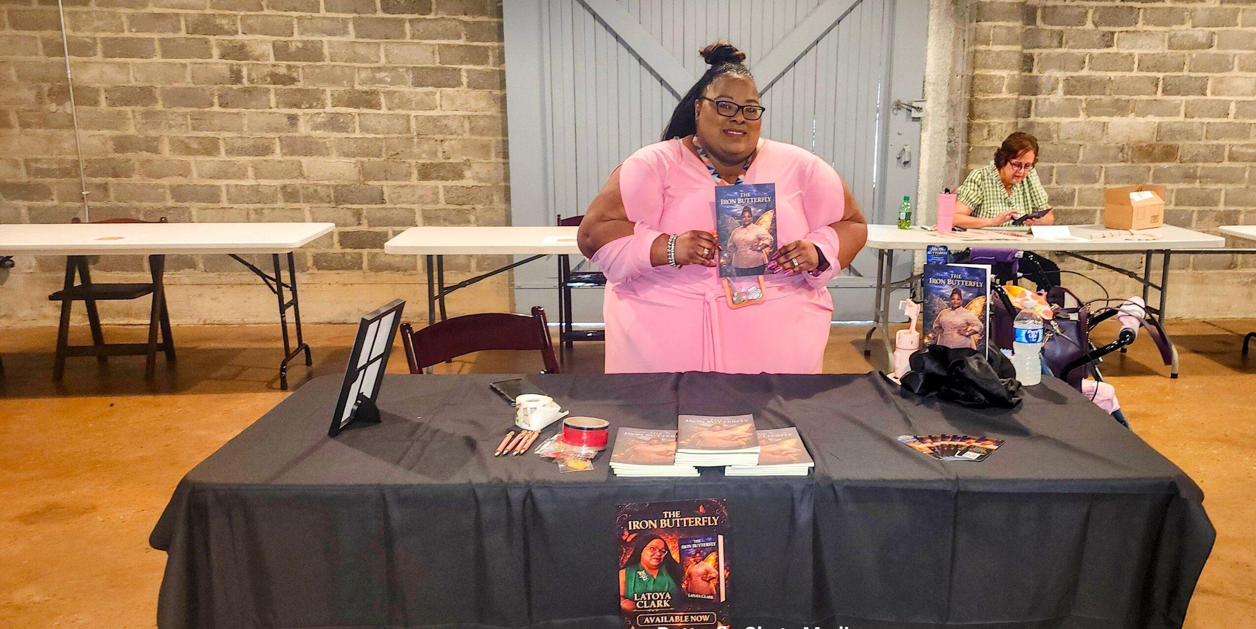 Person standing behind a table with books and merchandise, in a room with brick walls and other tables in the background.