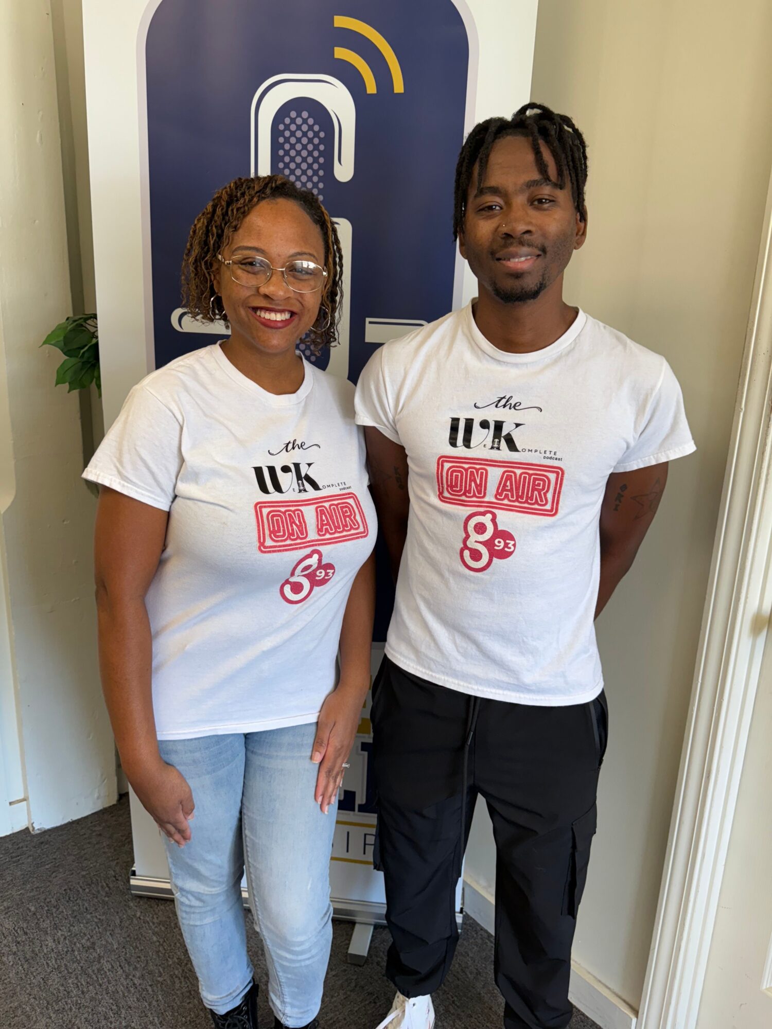 Two people standing indoors, one with braided hair and the other with short hair, both wearing white T-shirts with red and black text, smiling.