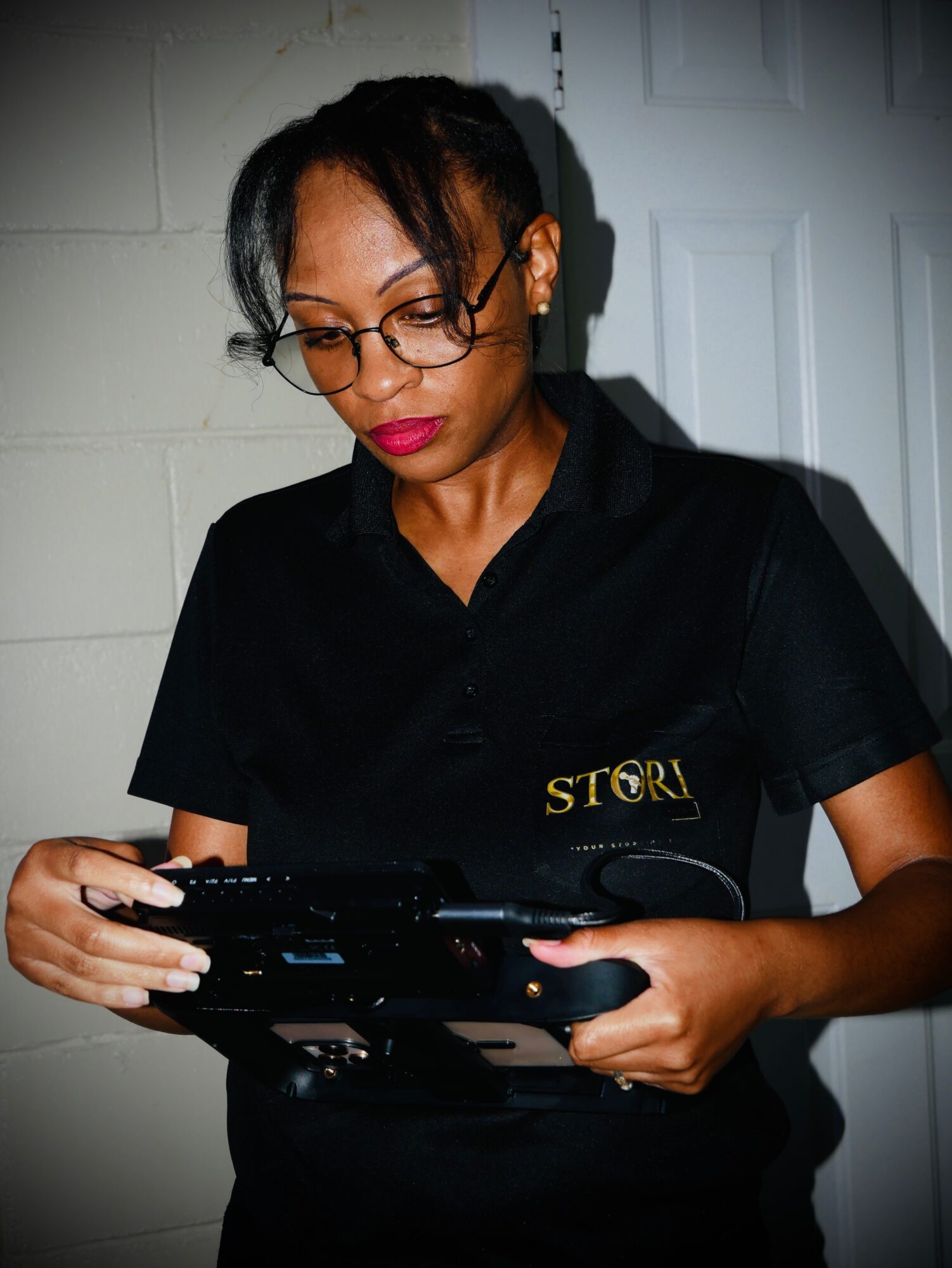 Woman with glasses in black uniform looking at a device, standing indoors near a white door.