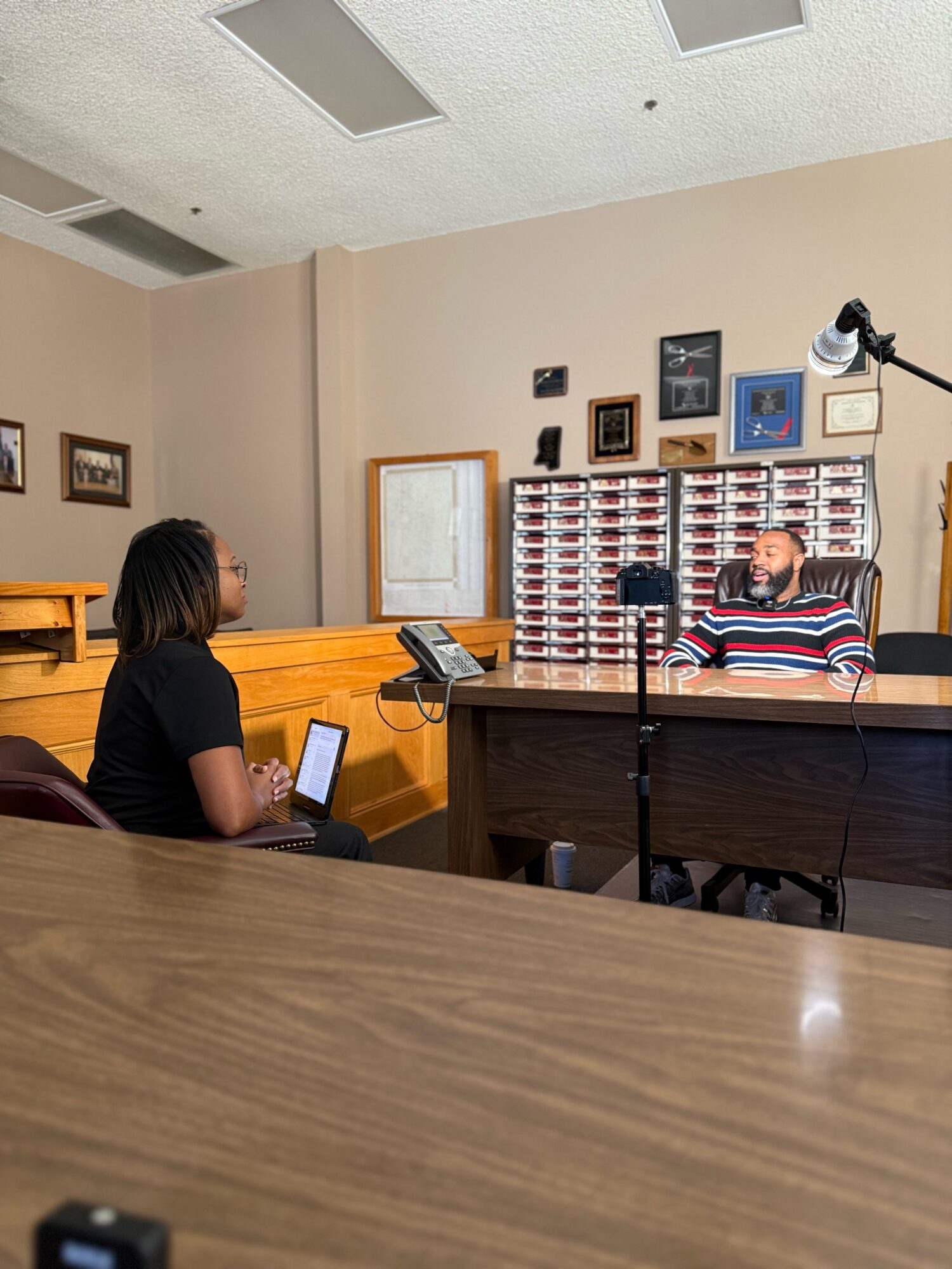 Two women in an office, one sitting at a desk and the other standing, with framed pictures and a cabinet behind the desk.