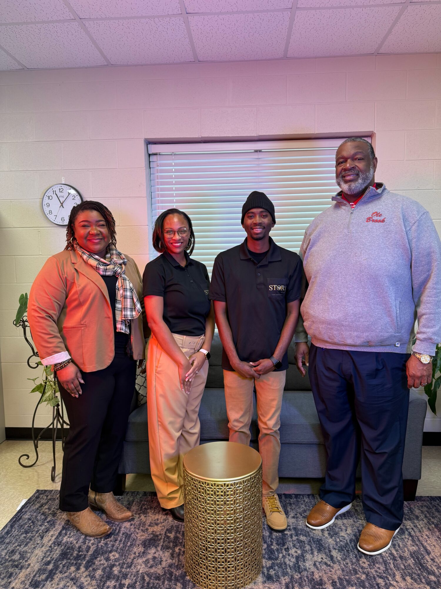 Four people standing indoors in front of a window with blinds, smiling, with a trash can in front, and a clock on the wall.