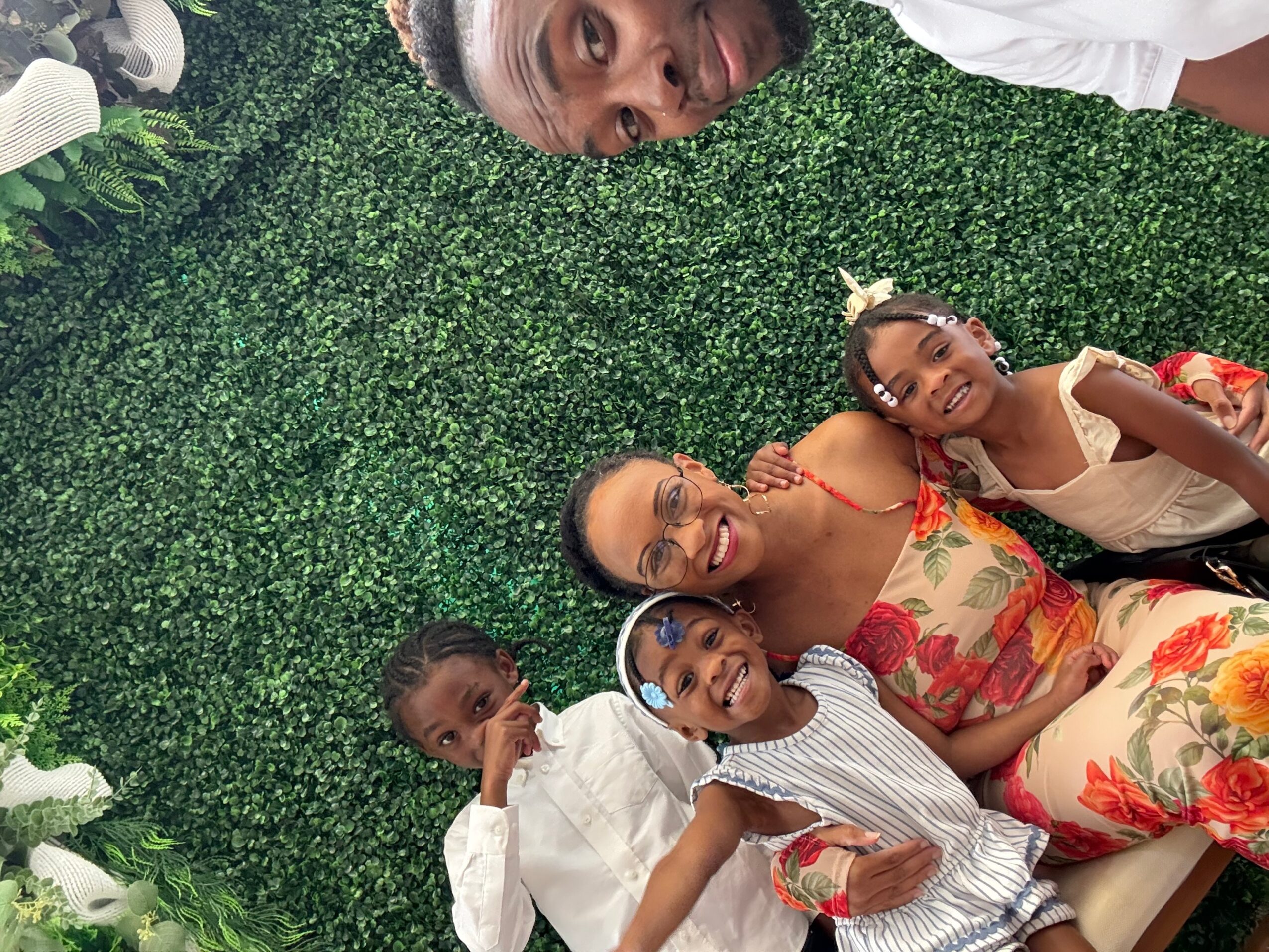 Four people lying on green grass, smiling and looking up at the camera, with one person at the top and three below.