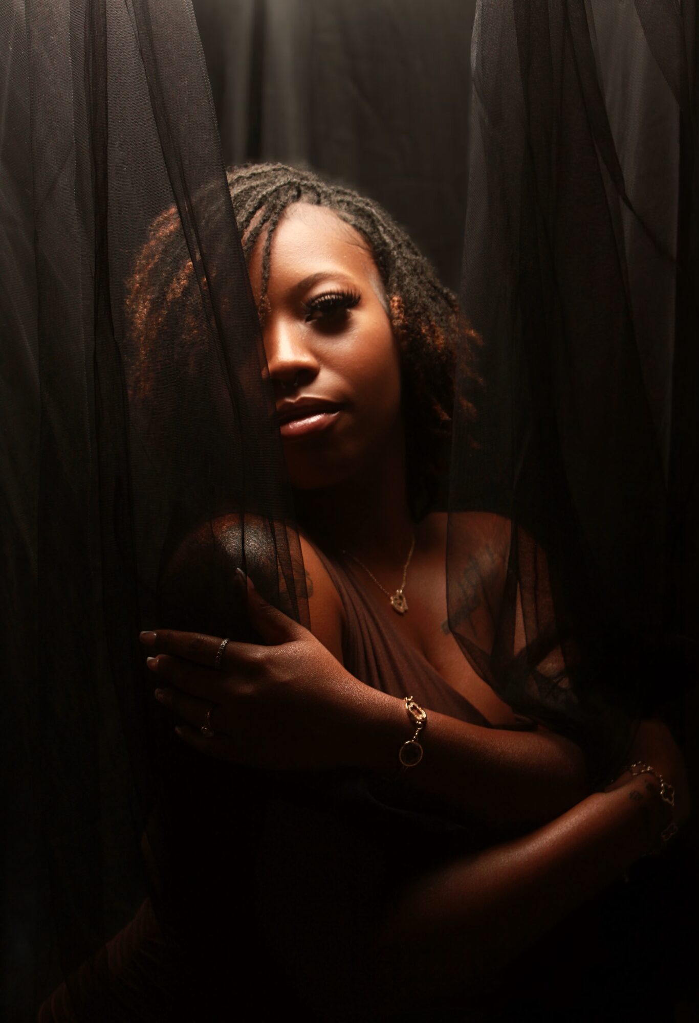 Portrait of a woman with braided hair, partially obscured by black fabric, looking directly at the camera, holding her arm.