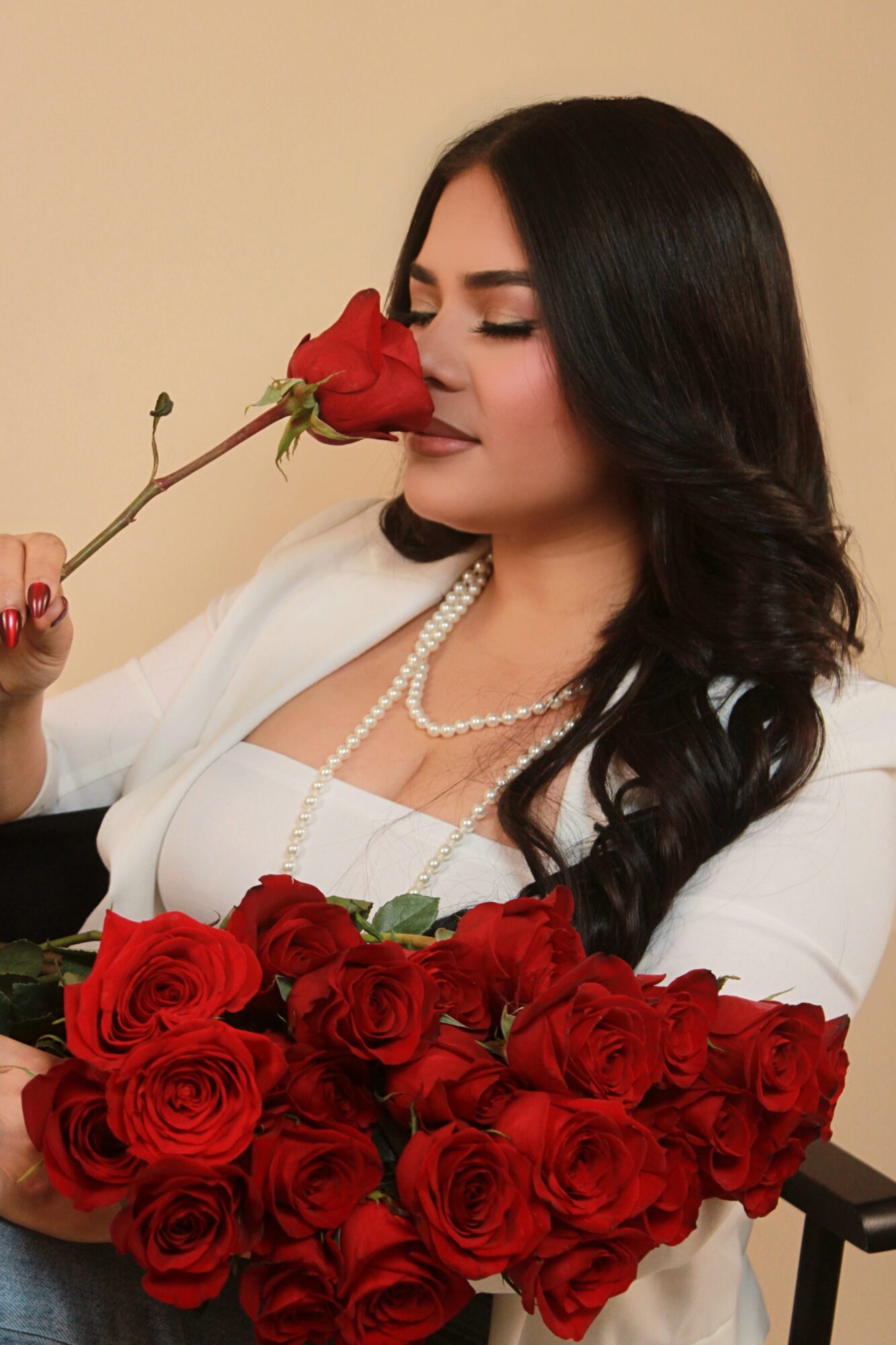 Woman with long dark hair holding a red rose near her face, surrounded by a bouquet of roses, wearing a white outfit and pearl necklace.