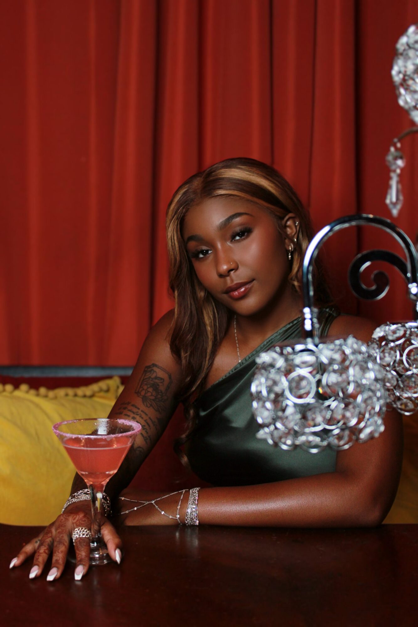 Woman with long hair sitting at a table with a pink cocktail, red curtains in background, elegant jewelry, and a crystal chandelier.