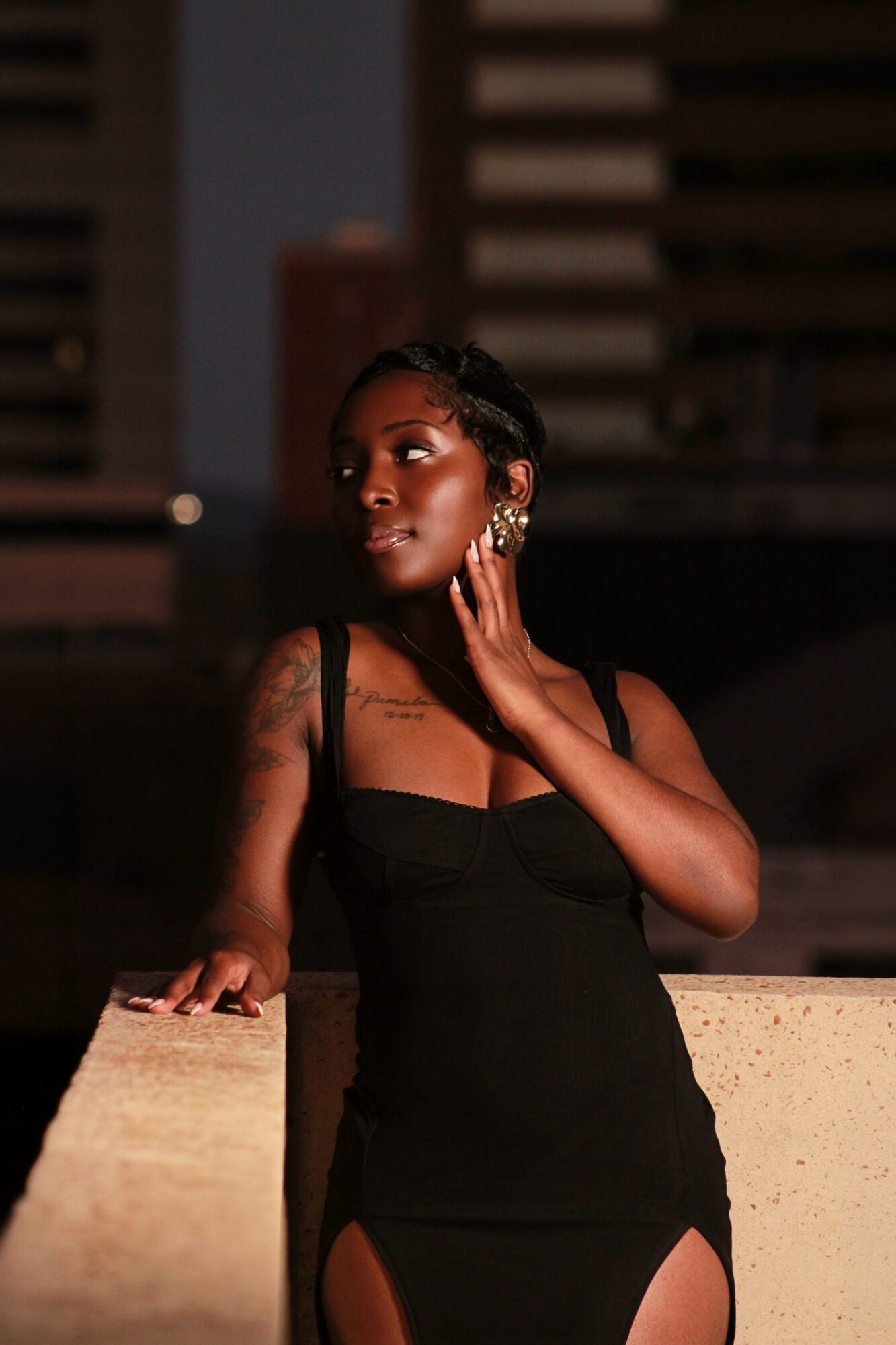 Woman in black dress with tattoos, standing outdoors at night, touching her face, city buildings in background.