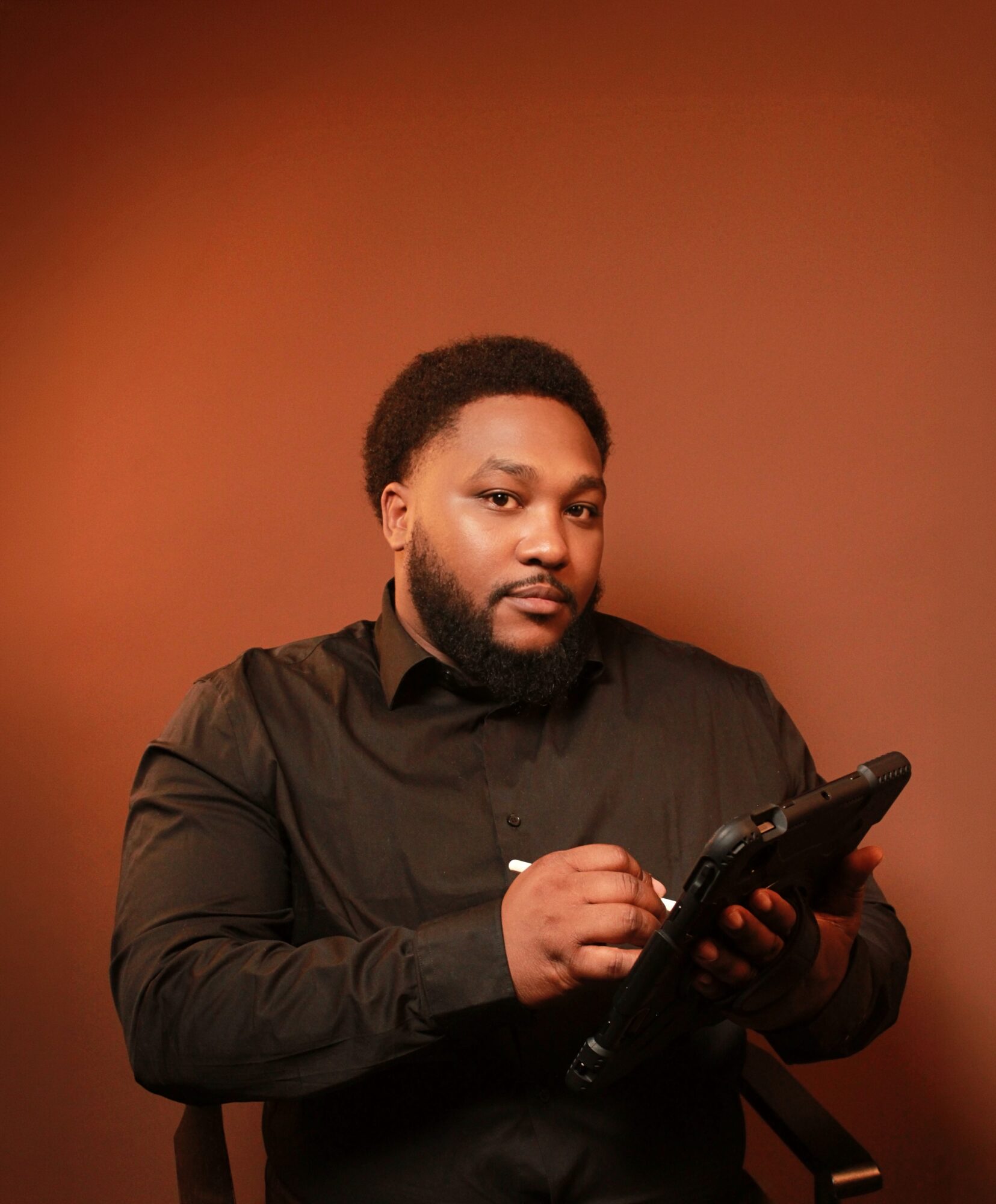 Man with a beard and short hair wearing a black shirt, holding a tablet, against a brown background.
