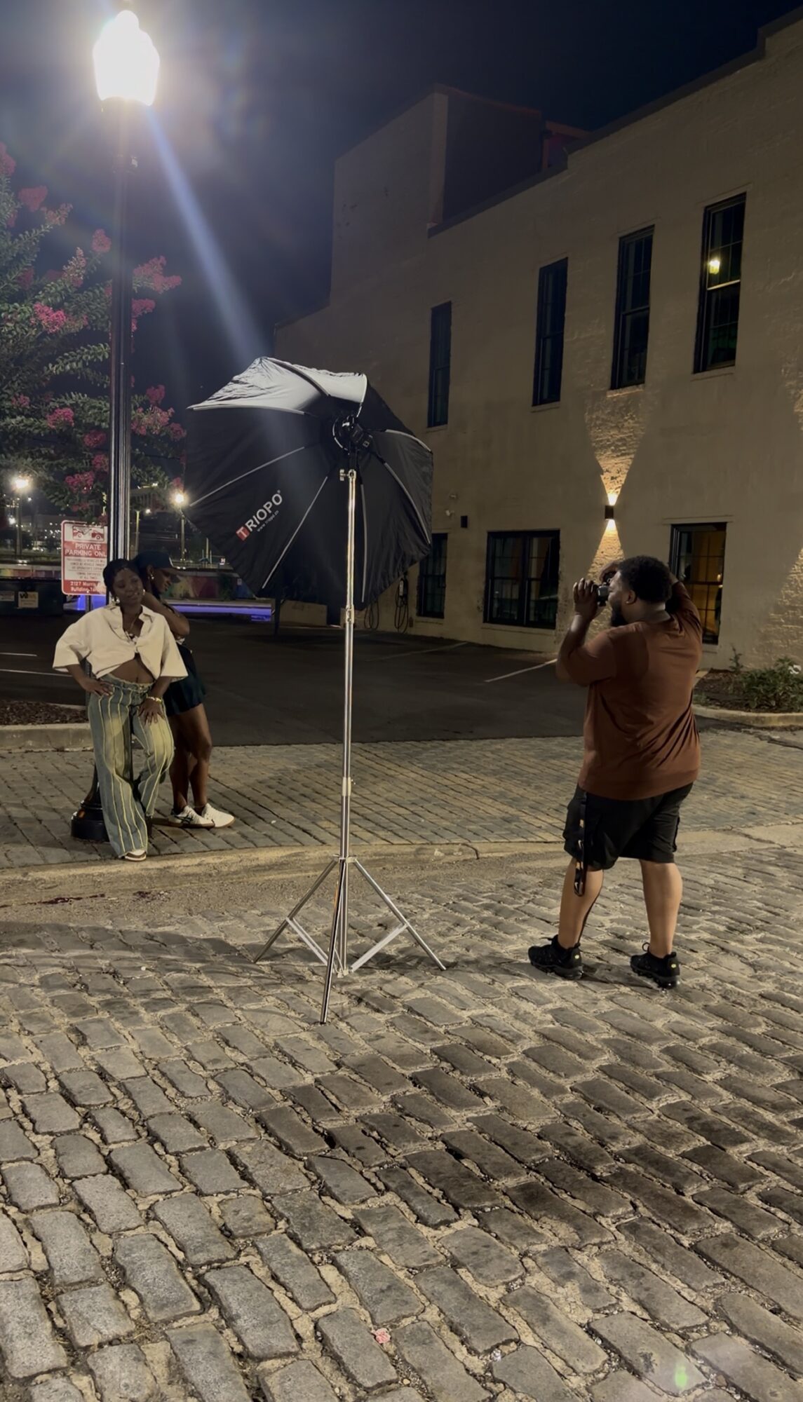 Person taking a photo with a camera on cobblestone street at night, with a building and streetlamp in background.