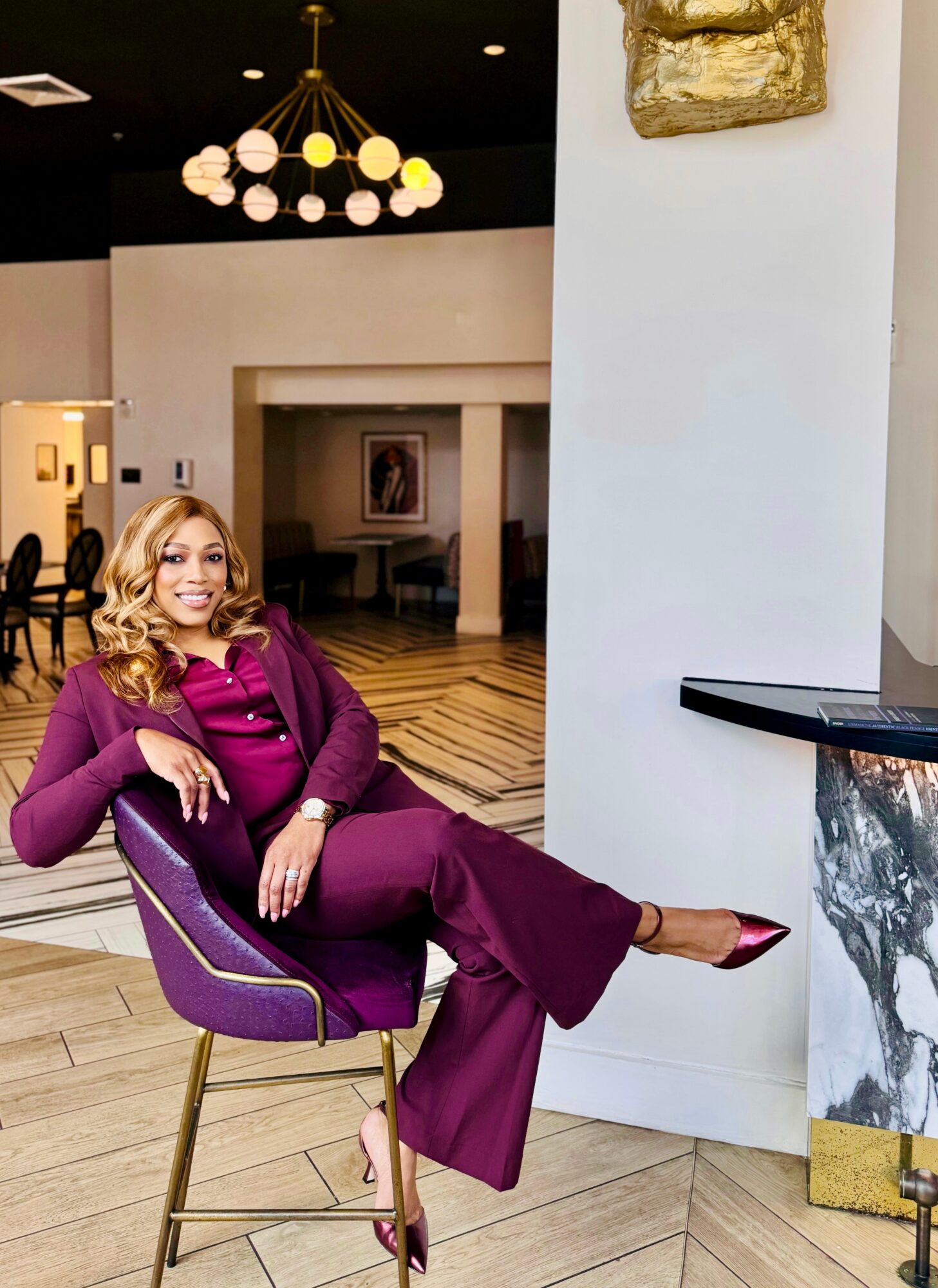 Woman in purple outfit sitting on a purple chair in a modern interior with a chandelier and artwork.