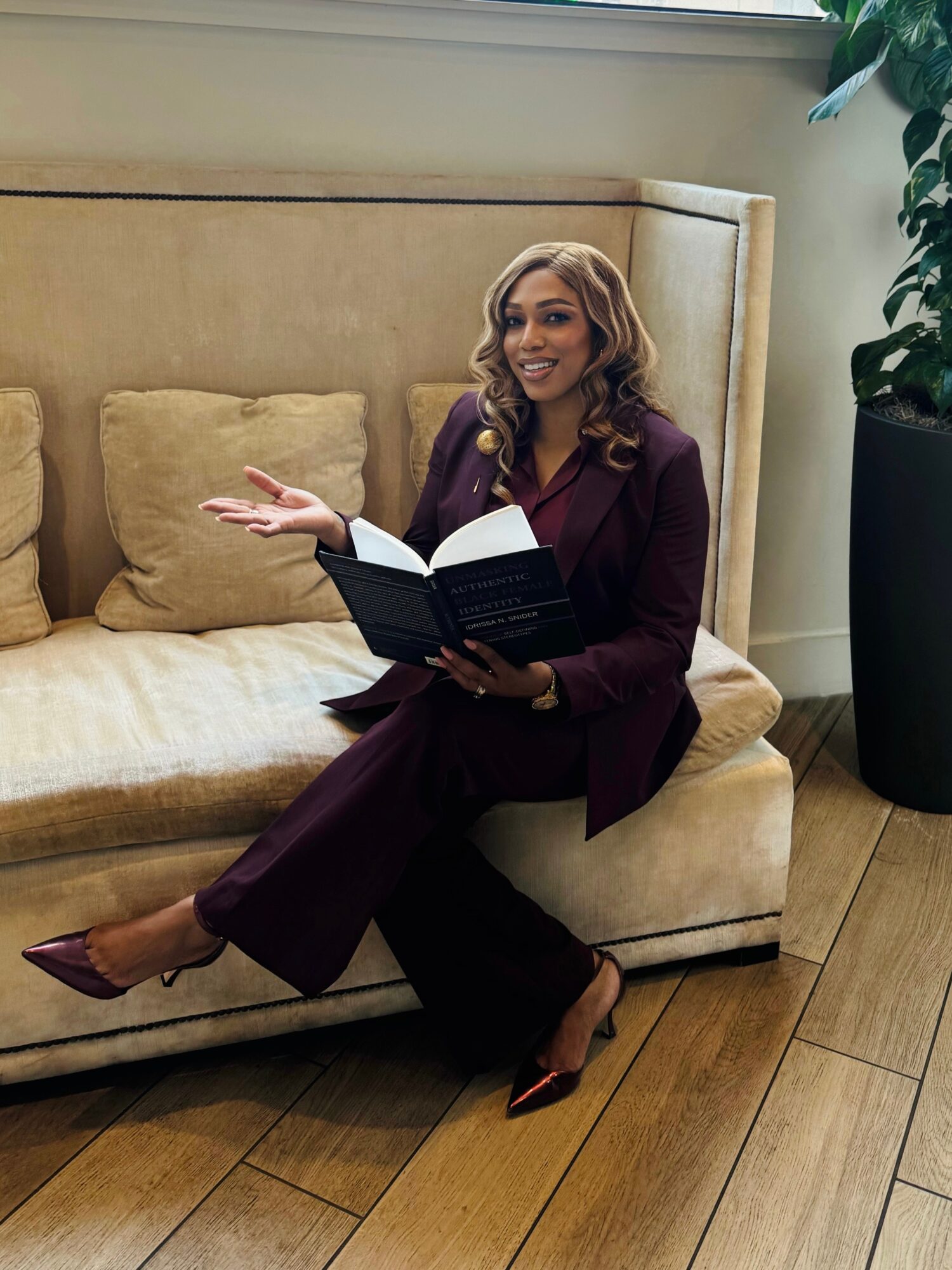 Woman sitting on a beige sofa holding a book, smiling, with a large potted plant nearby.