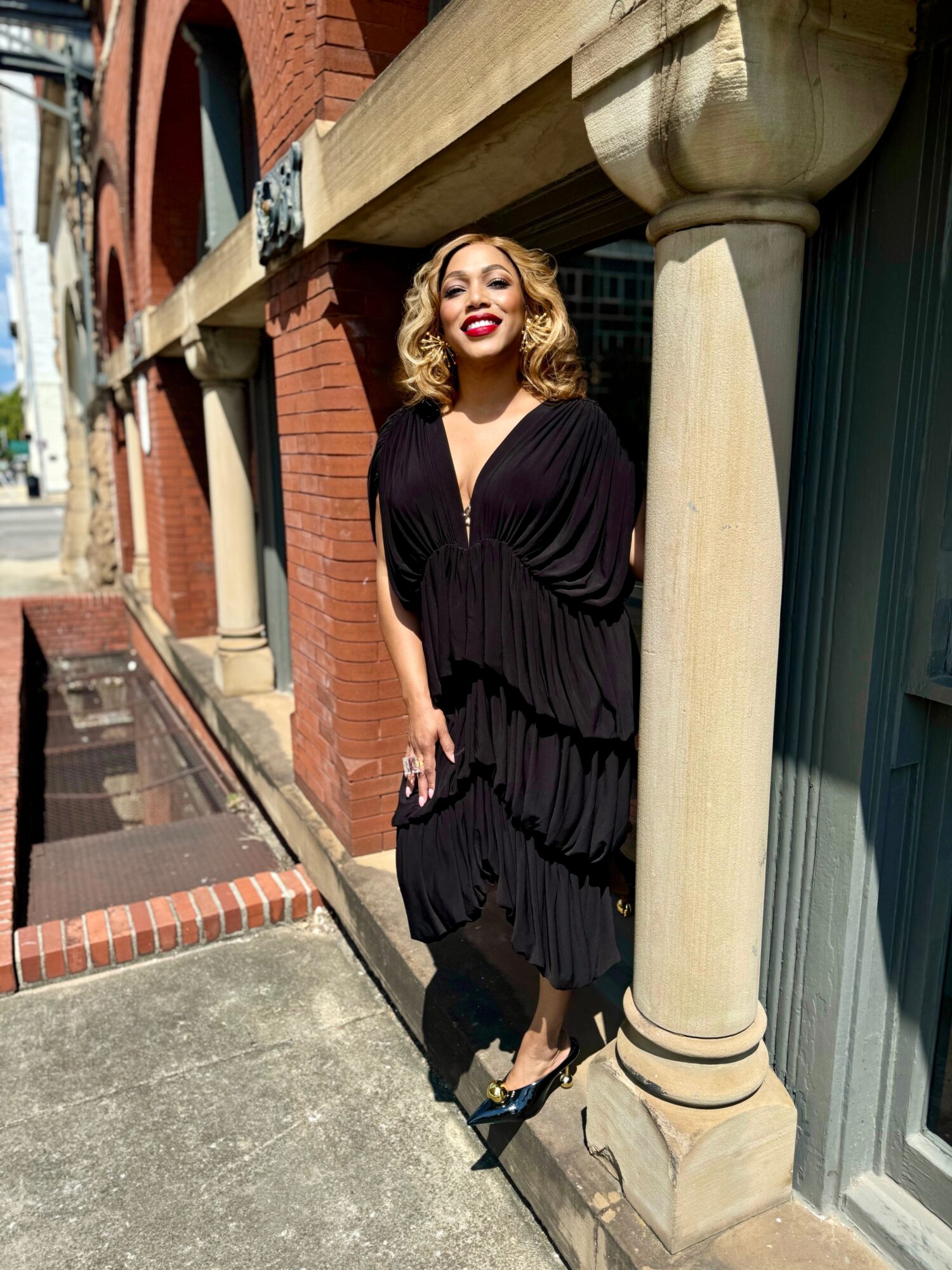 Woman in black dress standing outdoors next to a column, smiling, with brick and building background.