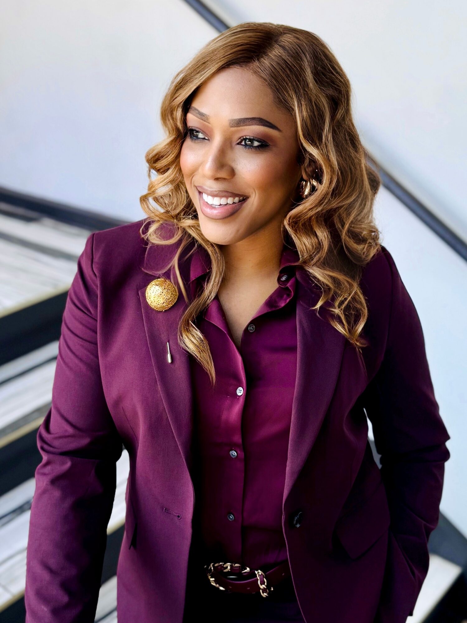 Woman with wavy hair smiling, wearing a purple blazer and shirt, standing indoors.