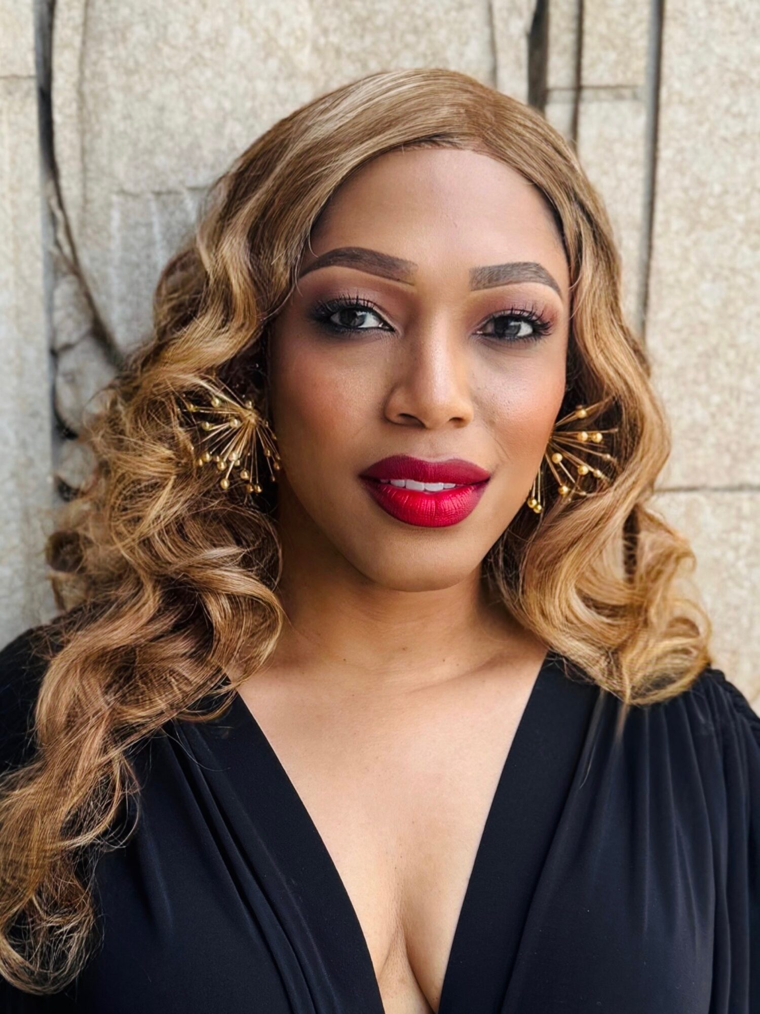 Woman with wavy blonde hair, red lipstick, and gold earrings, wearing a black dress, standing outdoors against a stone wall.
