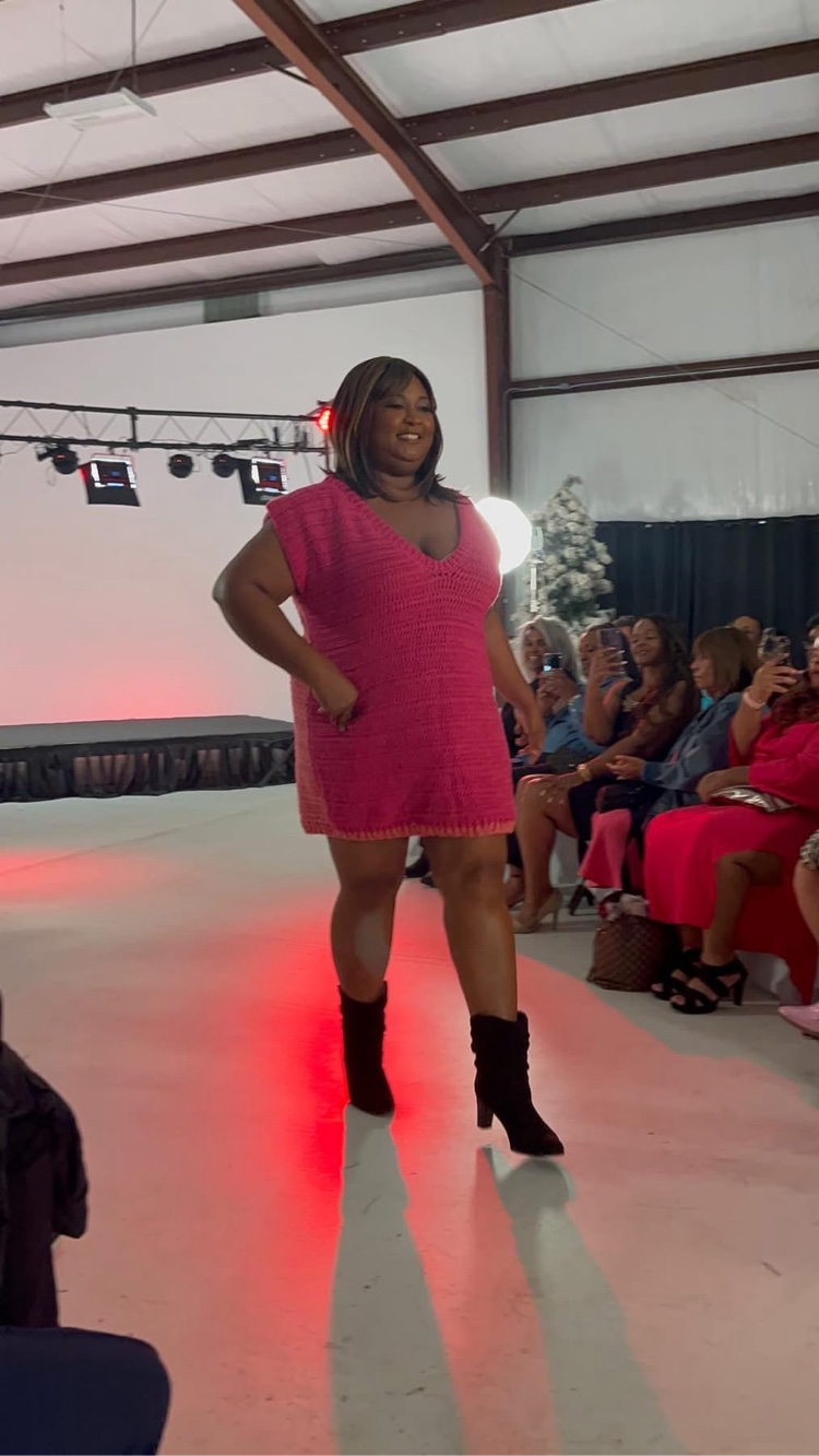 Woman walking on runway in pink dress and black boots, audience seated on right side, industrial ceiling above.