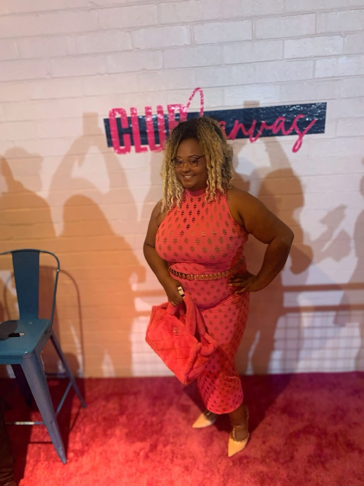 Woman in a red dress standing on a red carpet, smiling, with a white brick wall background and a sign behind her.