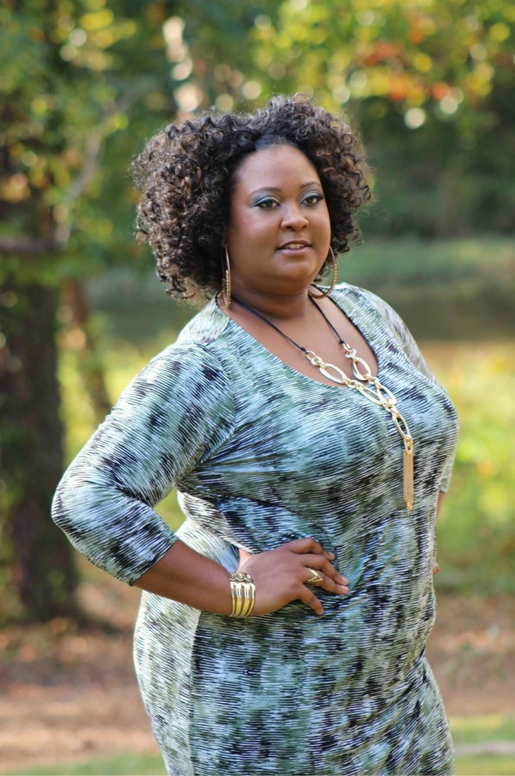 Woman with curly hair standing outdoors in a park, wearing a patterned dress and jewelry, with trees and greenery in background.