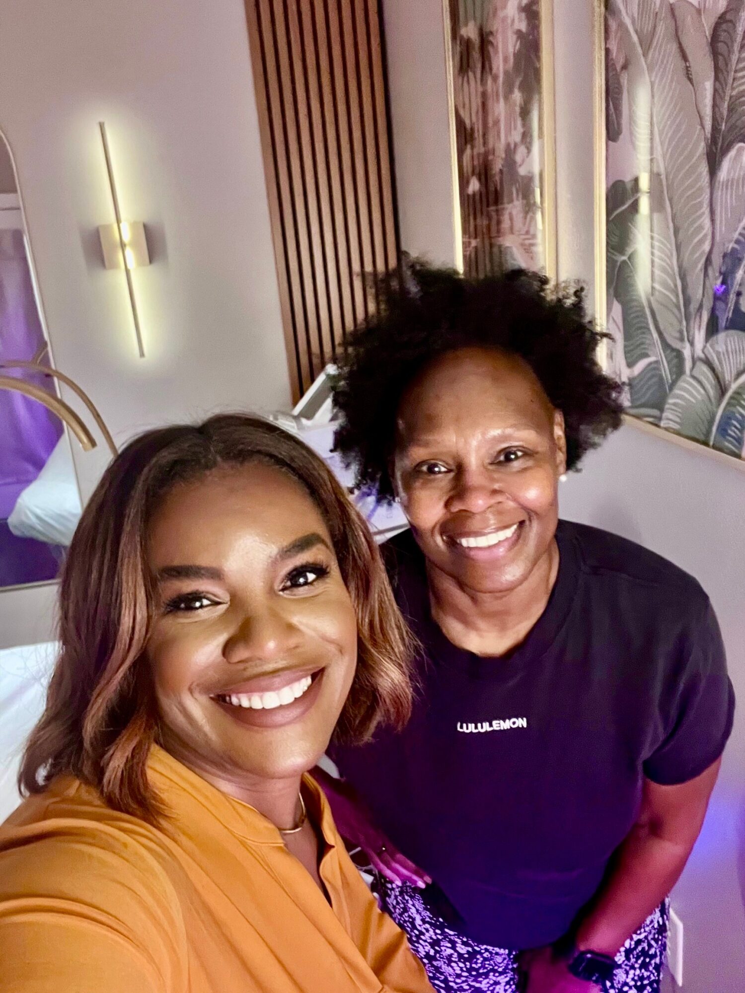 Two women smiling indoors, one with long hair and the other with short curly hair, standing close together.
