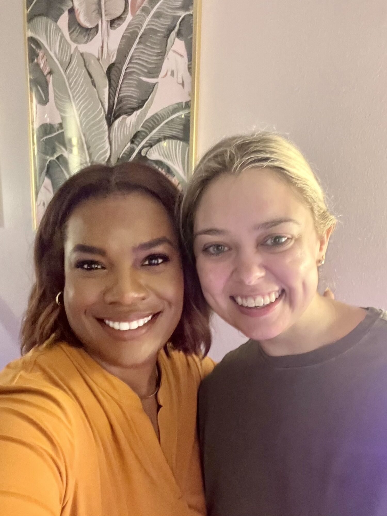 Two women smiling close together indoors, with a large botanical artwork in the background.