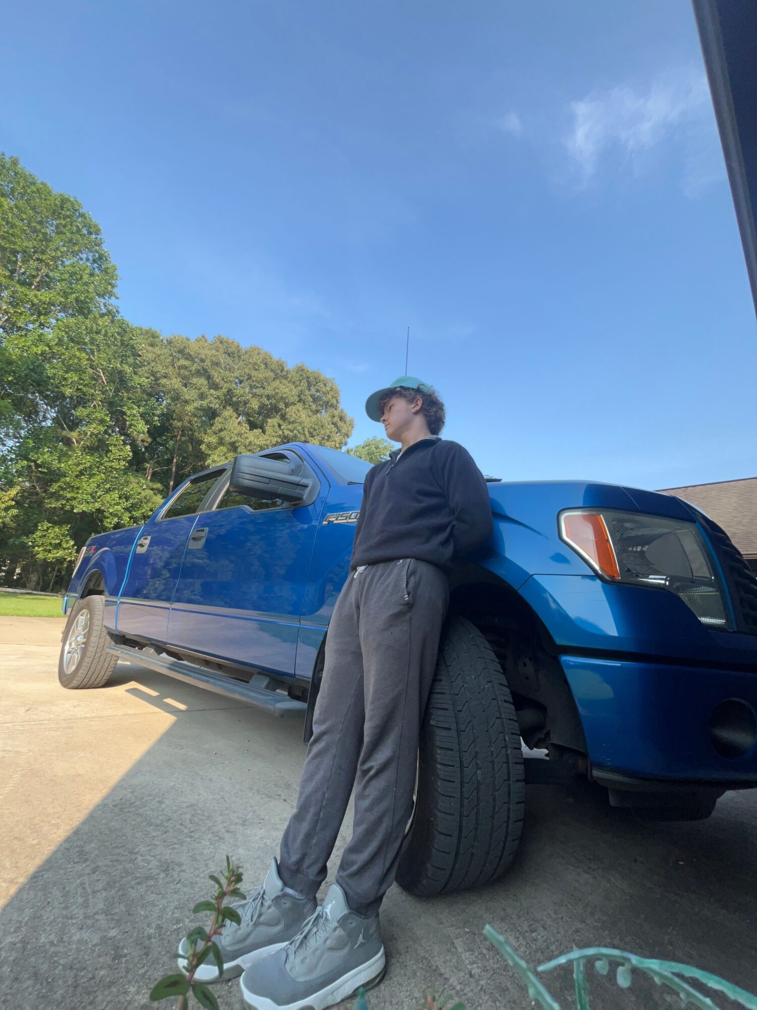 Person standing next to a blue car outdoors with trees and a clear sky in the background.