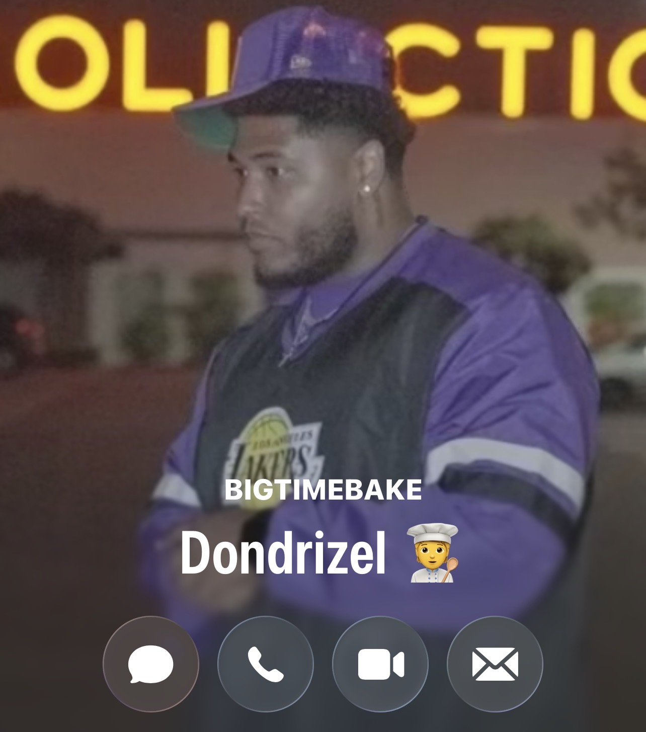 Young man in purple and black jacket and cap, standing outdoors at night, with a sign in the background.