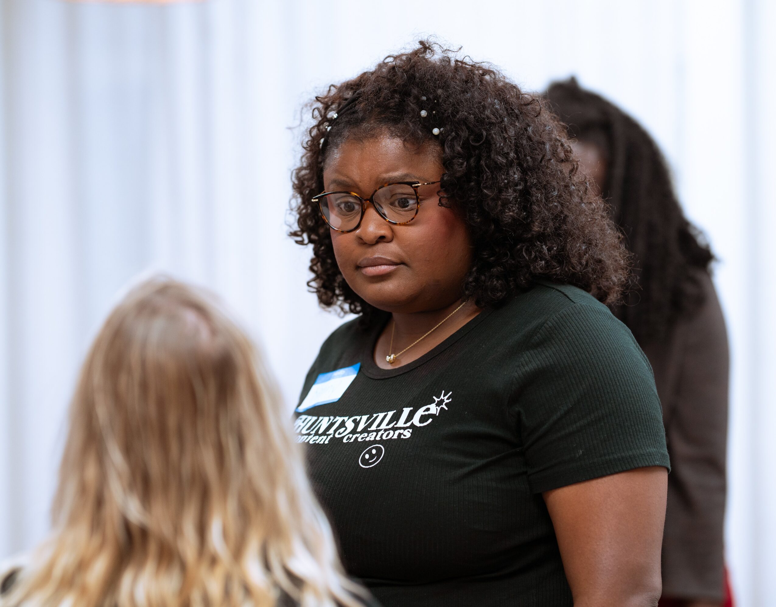 Woman with curly hair and glasses looking at a person with blonde hair in a room with light curtains.