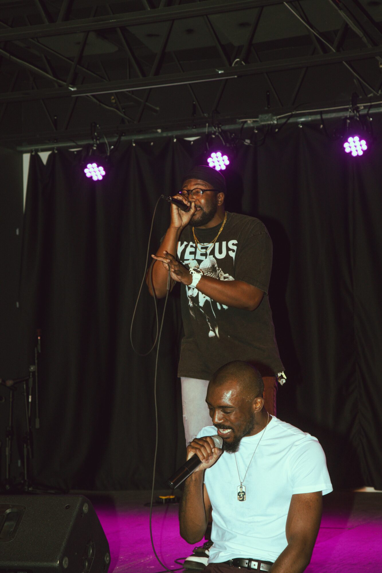 Two men perform on stage with black curtains and purple stage lights, one singing into a microphone, the other with a microphone in hand.