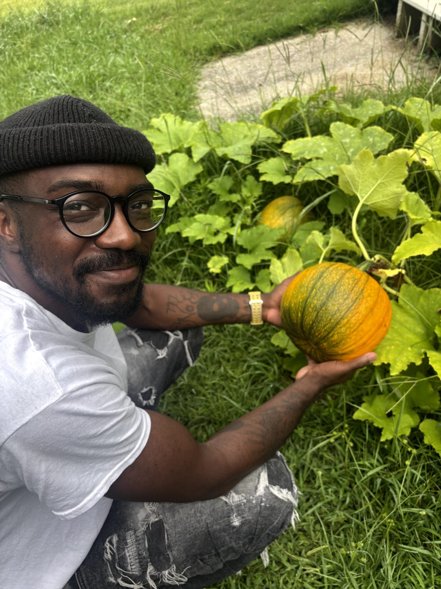 Man with glasses and a black beanie holding a pumpkin in a garden with green leaves and grass