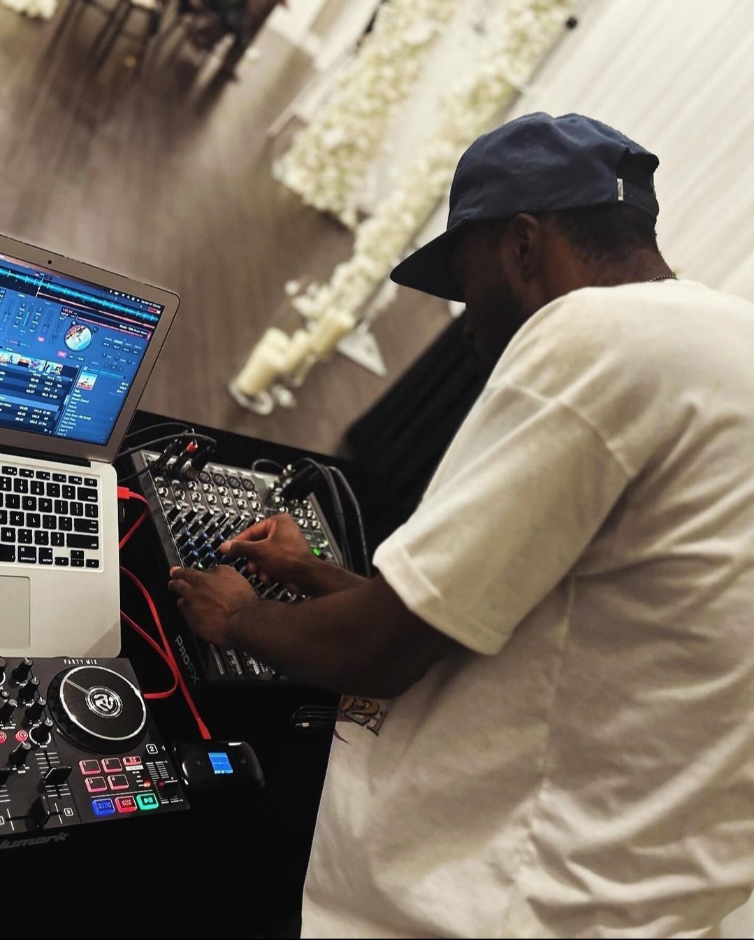 Person wearing a cap and white shirt operating DJ equipment with a laptop nearby, in a decorated venue.