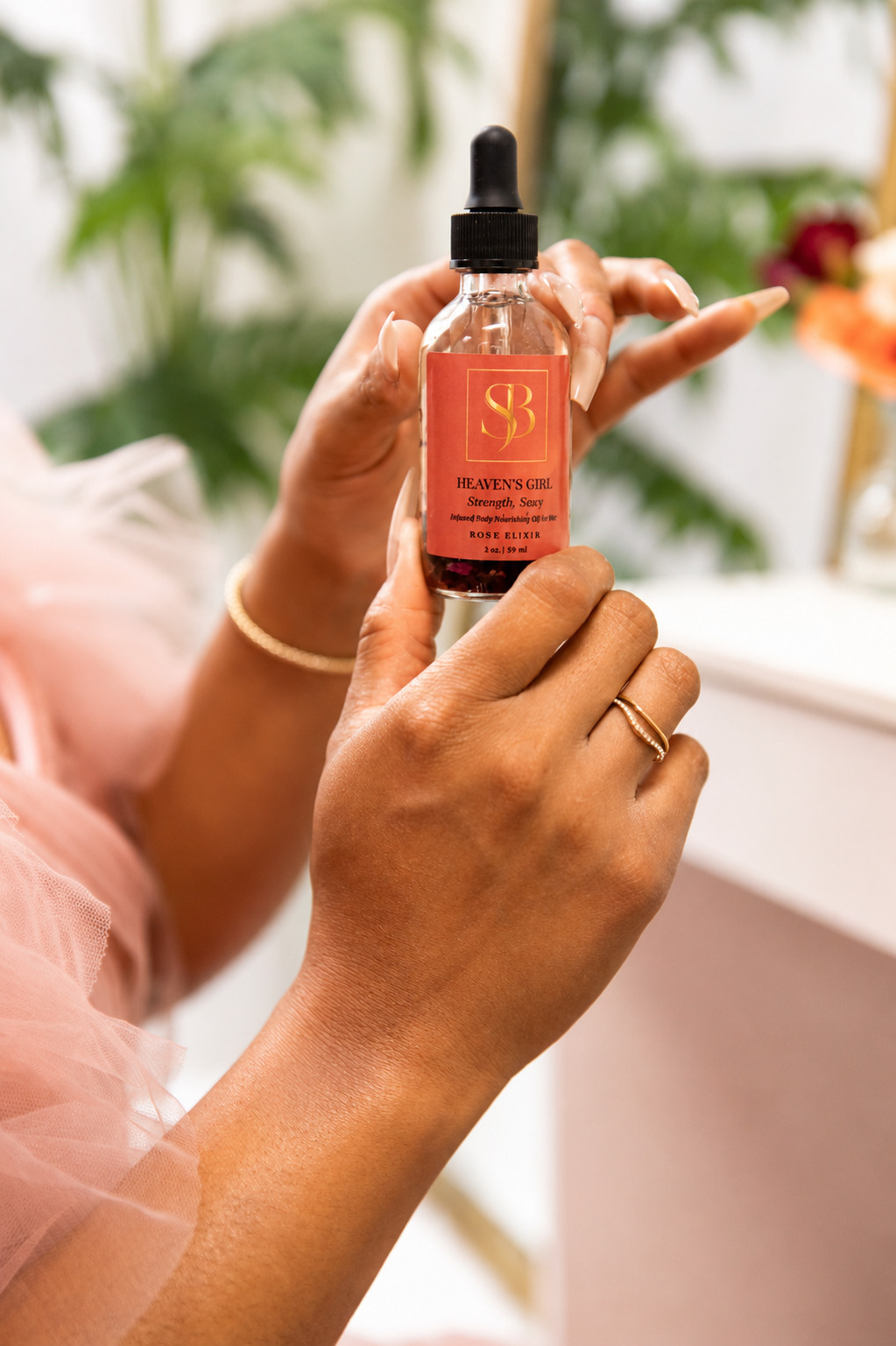Person holding a small glass spray bottle with a pink label, indoors with plants in the background.
