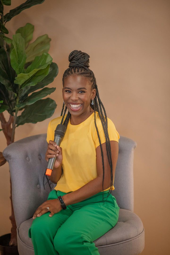 Smiling woman with braided hair in a bun, holding a microphone, sitting on a gray chair, wearing a yellow top and green pants.