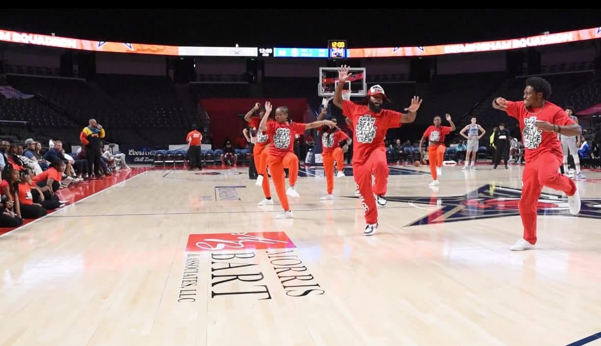 Group of dancers performing on a basketball court, some with arms raised, audience seated around the court, arena setting.