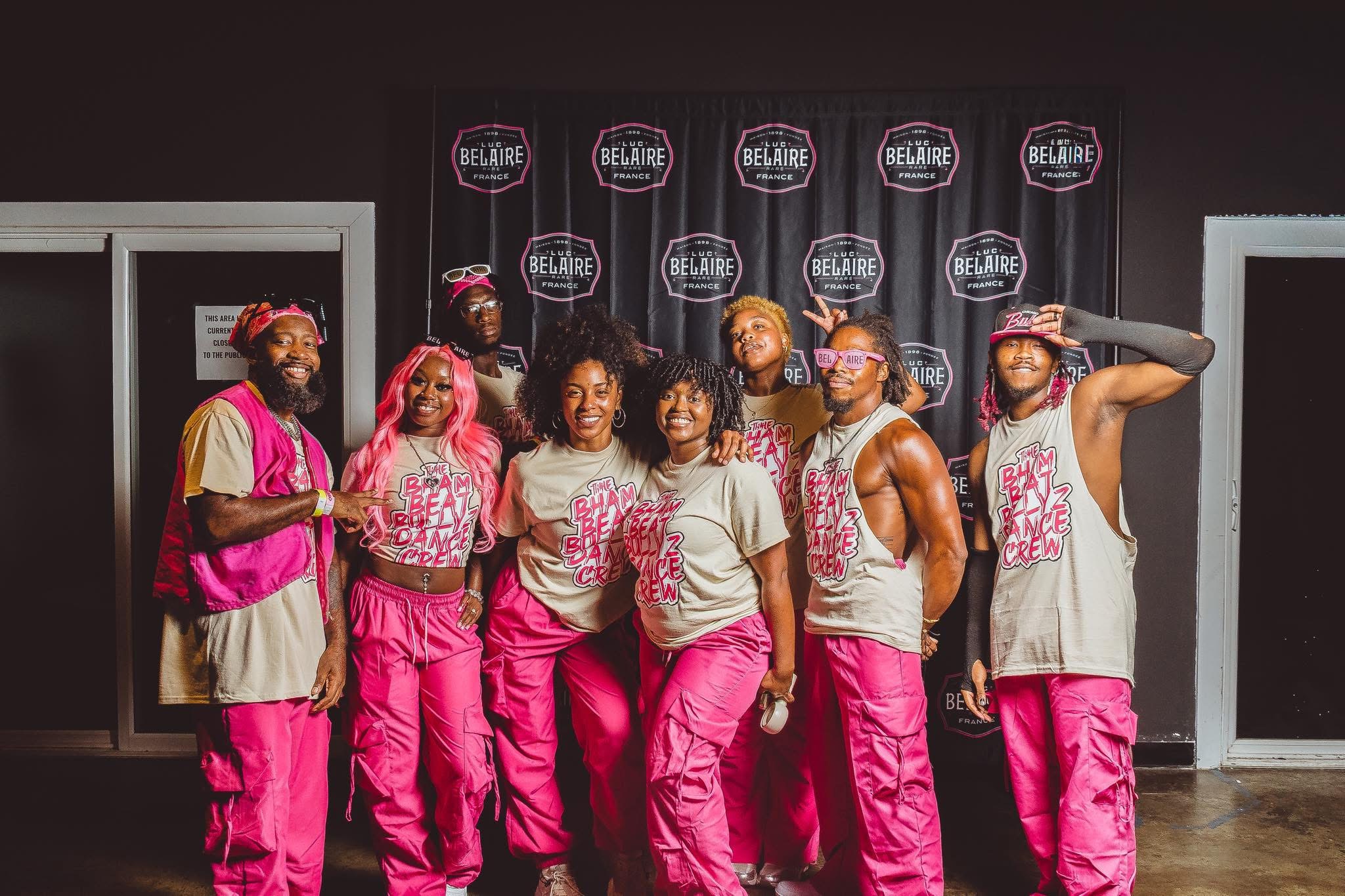 Group of seven people on stage wearing pink and white outfits, standing in front of a black backdrop with logos.