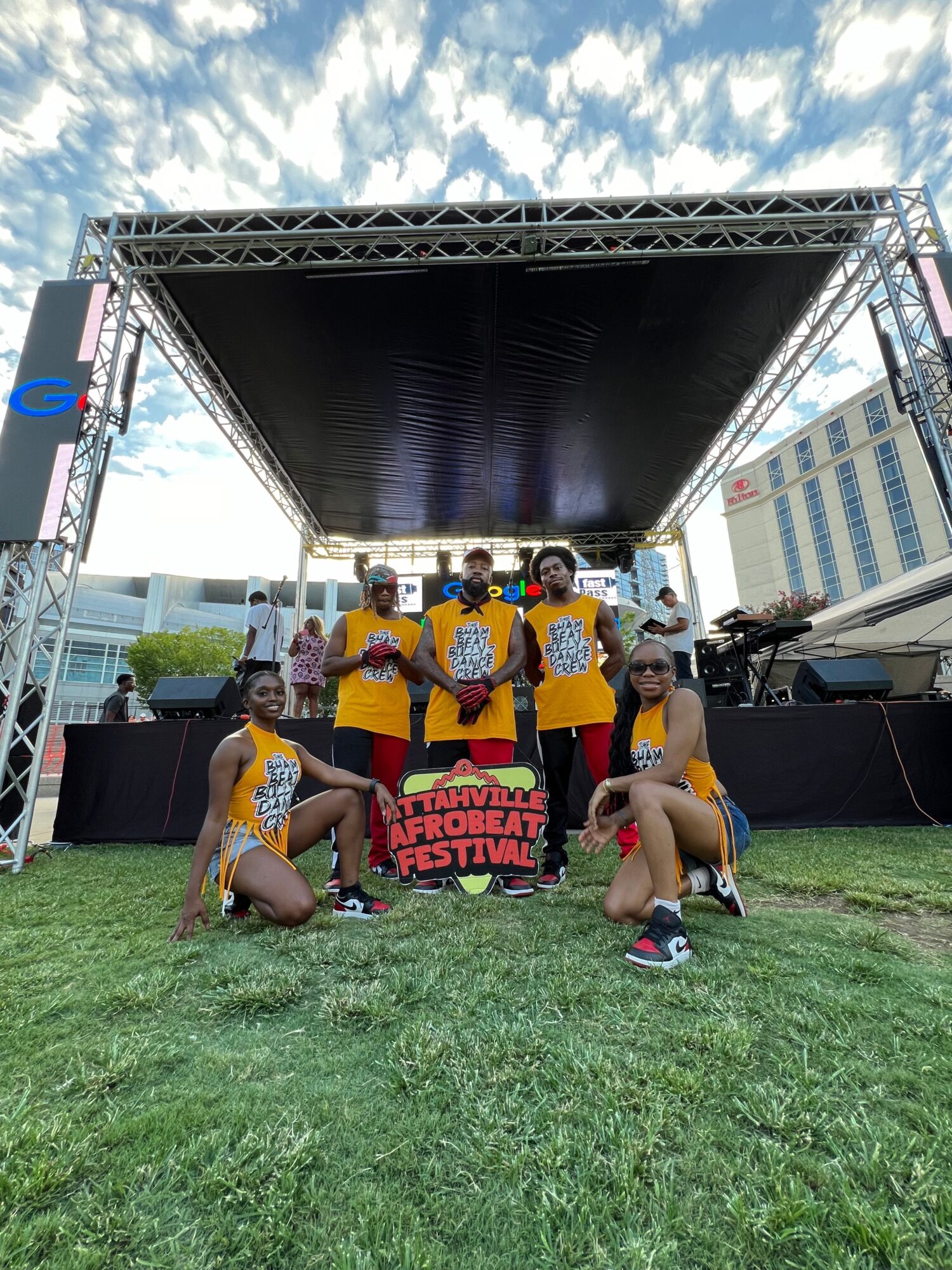 Group of children in yellow shirts and shorts gather on grass near a stage with a black backdrop, outdoors on a sunny day.