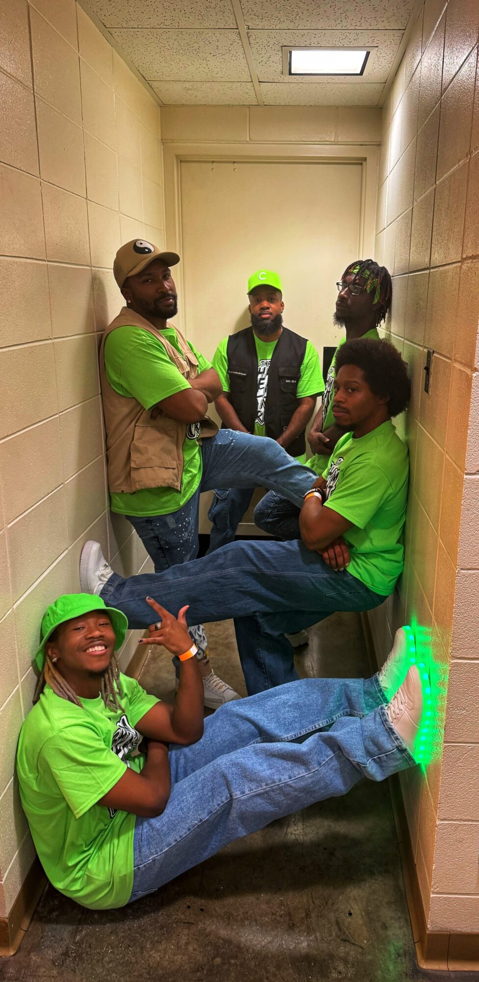 Five people in matching green shirts and hats sitting and standing in a narrow hallway, some smiling and posing.