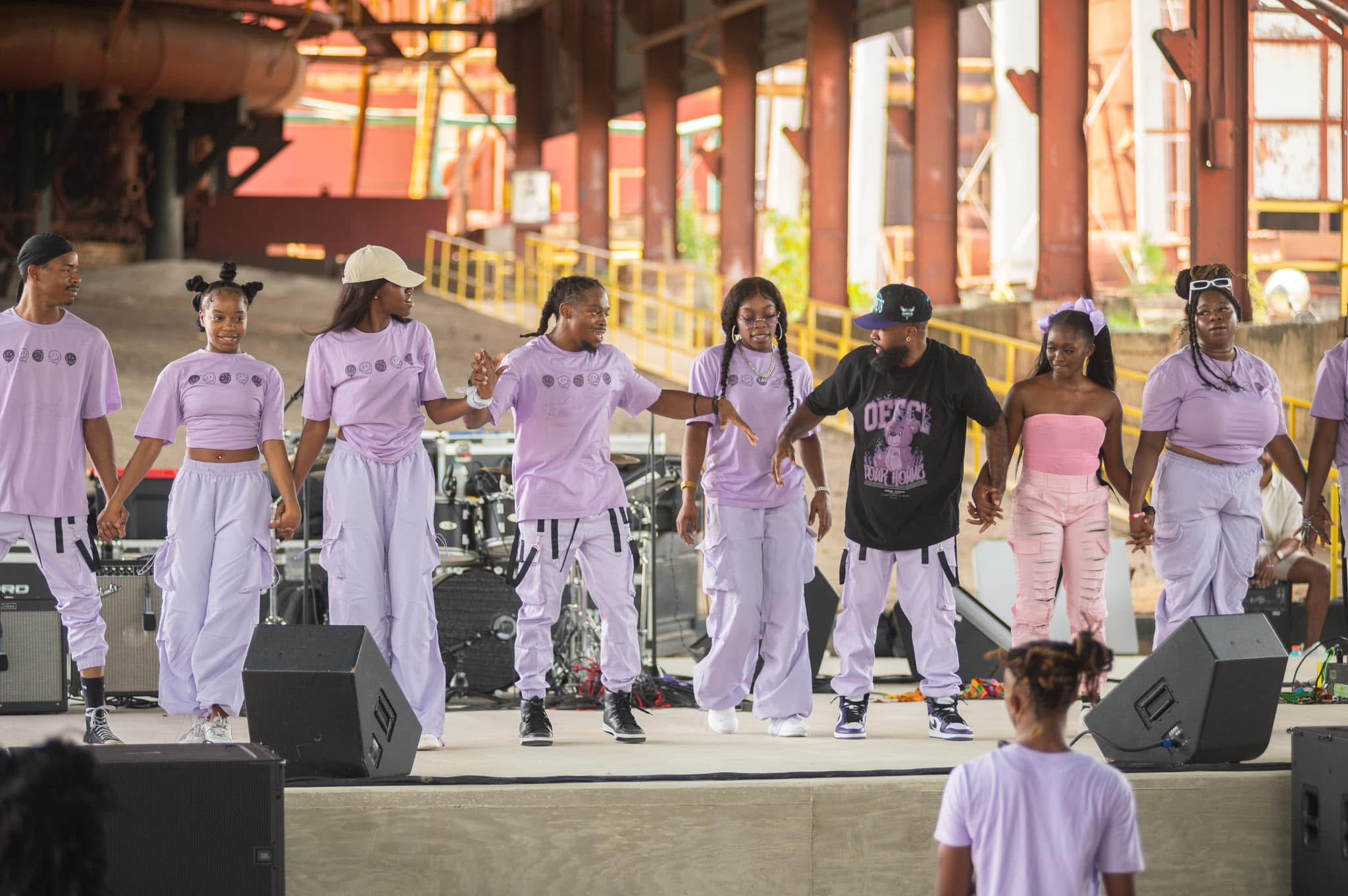Group of seven people performing on stage, some holding hands, in an industrial setting with large windows and equipment.