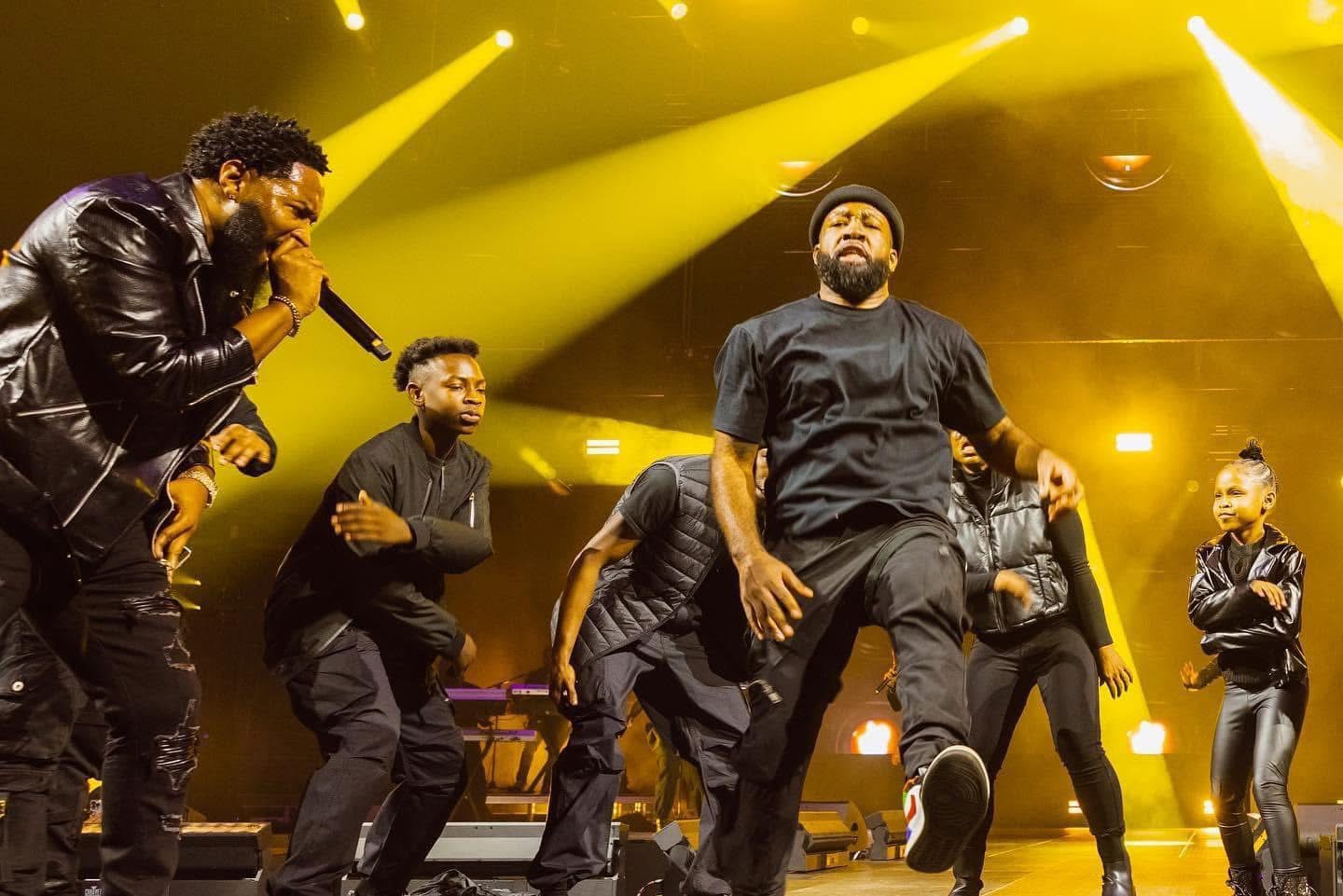 Group of six performers on stage with yellow lighting, singing and dancing, wearing black outfits.