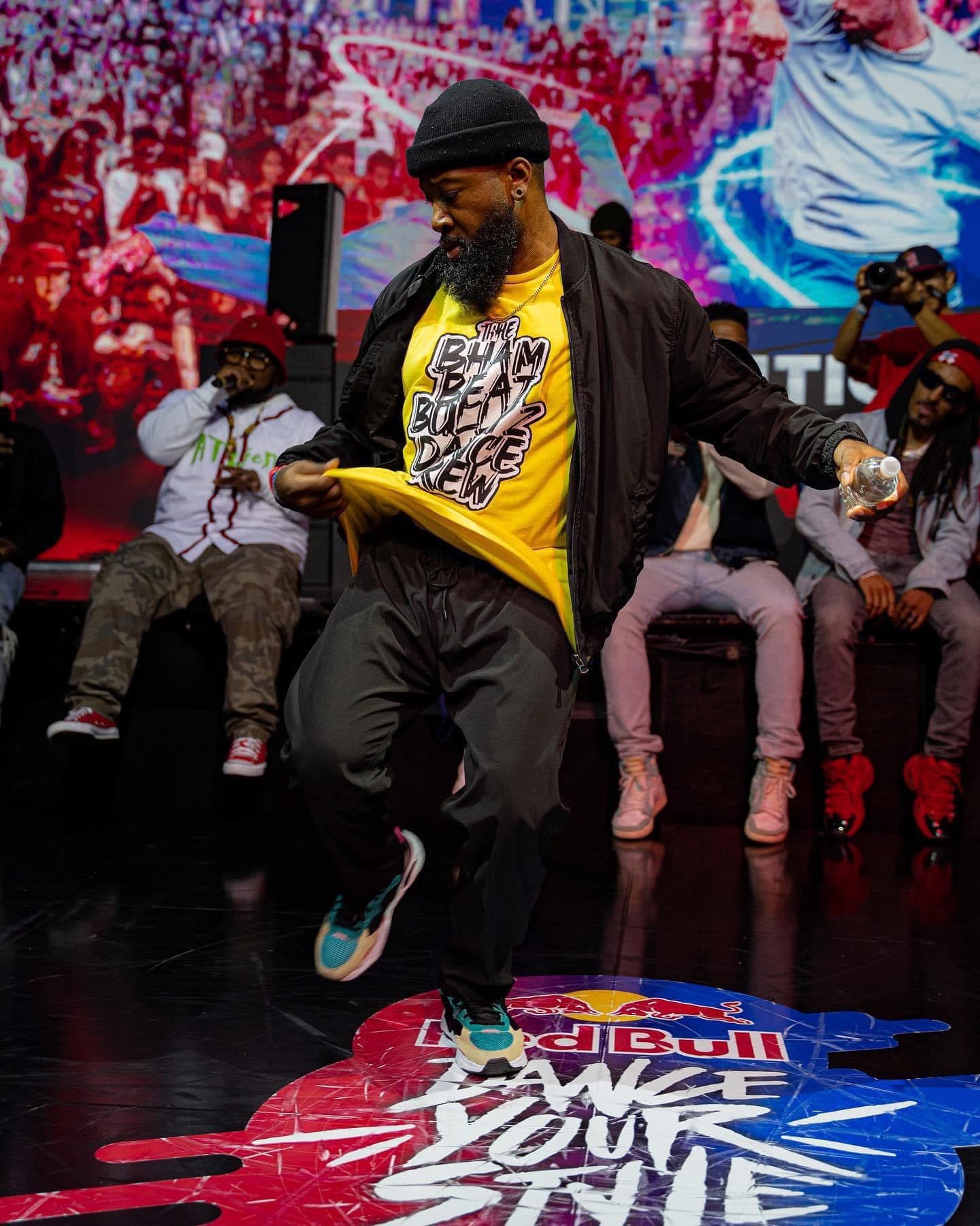 Man dancing on stage with a group of people, colorful background, Red Bull logo on stage floor, energetic scene.