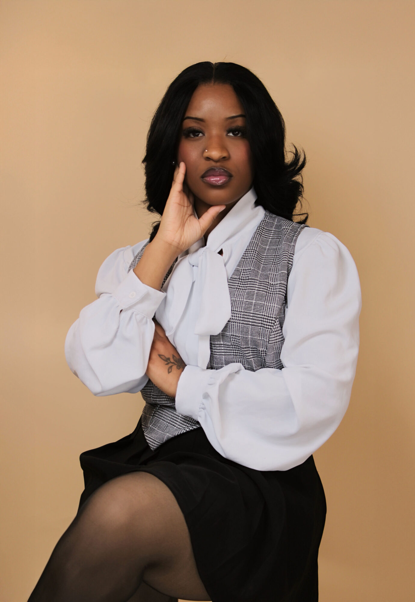 Young woman with black hair, wearing a white blouse with a bow, gray vest, and black skirt, sitting against a beige background.