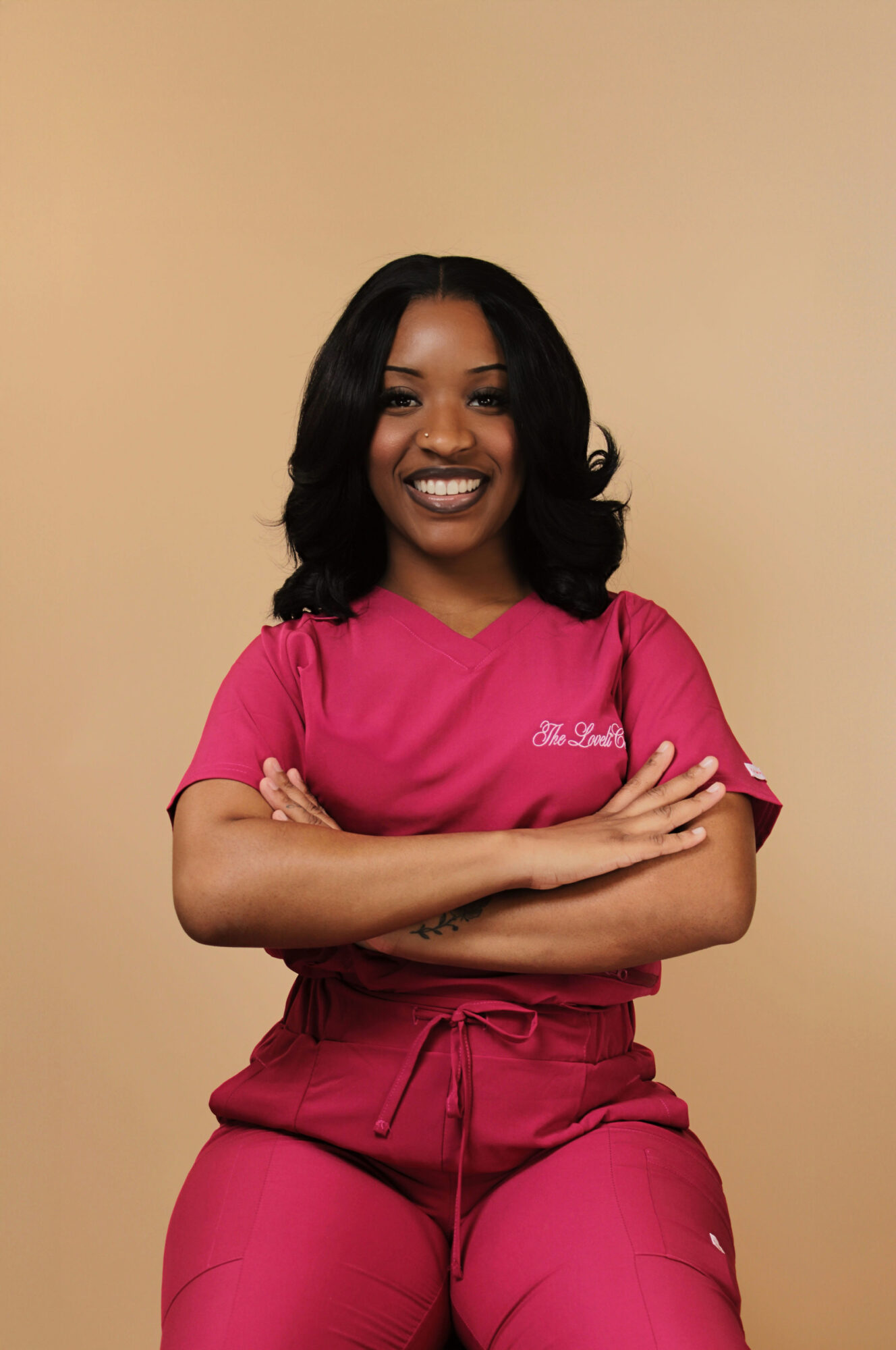 Smiling woman with black hair in pink scrubs, arms crossed, standing against beige background.