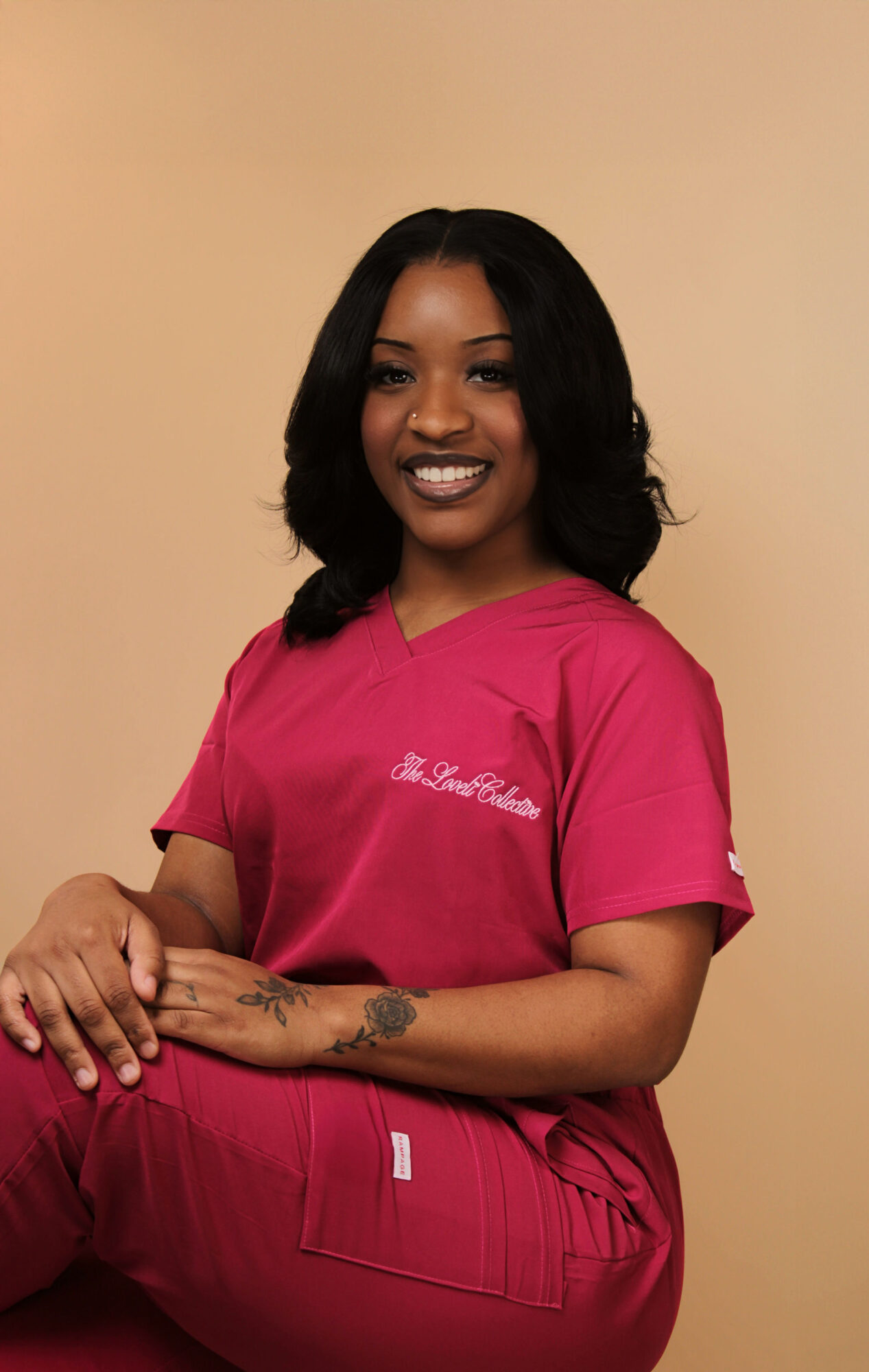 Young woman with shoulder-length black hair smiling, wearing a pink shirt and pants, standing against a beige background.