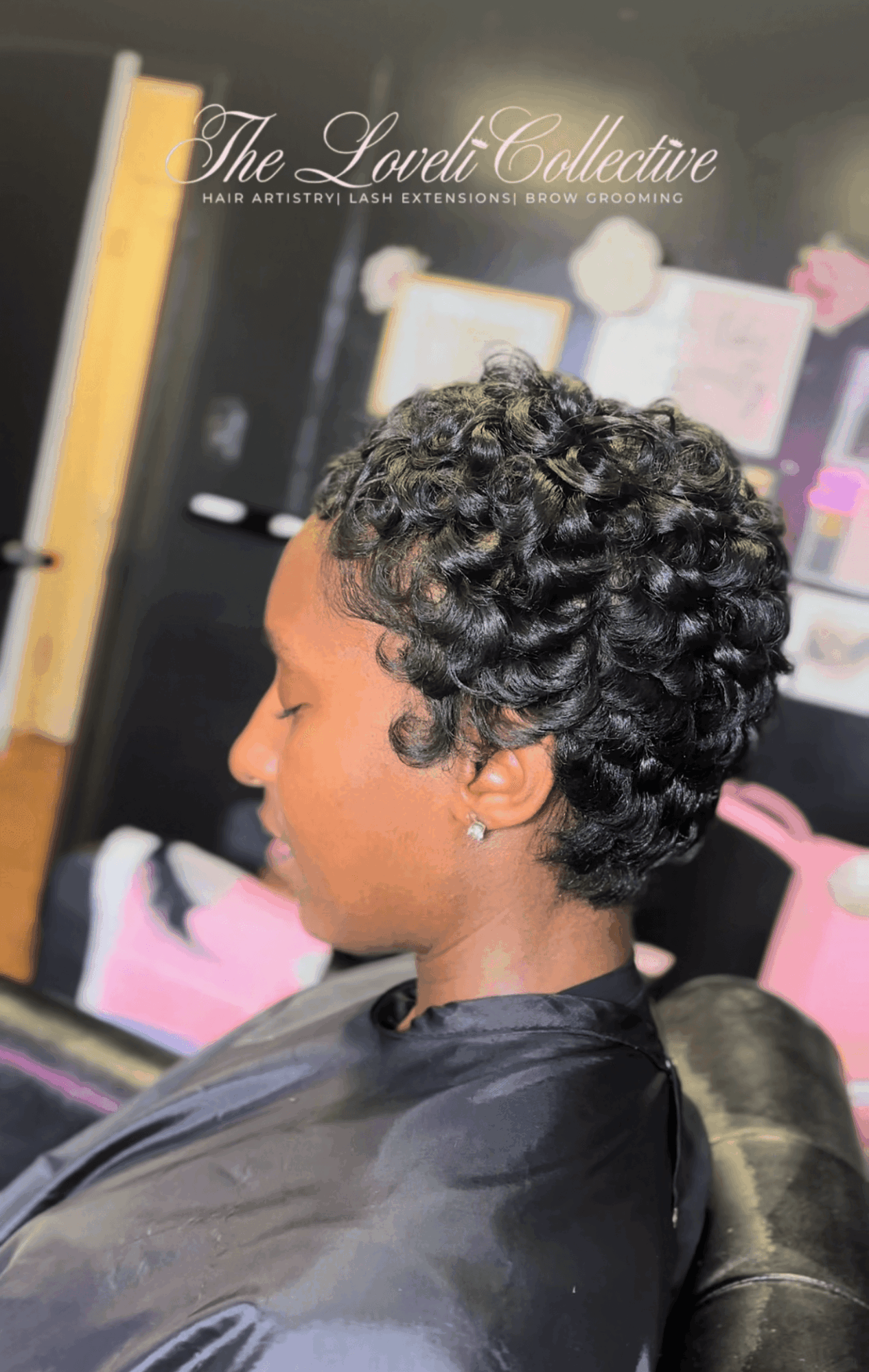 Side profile of a woman with short, curly black hair in a salon chair, with a blurred background of salon decor and products.