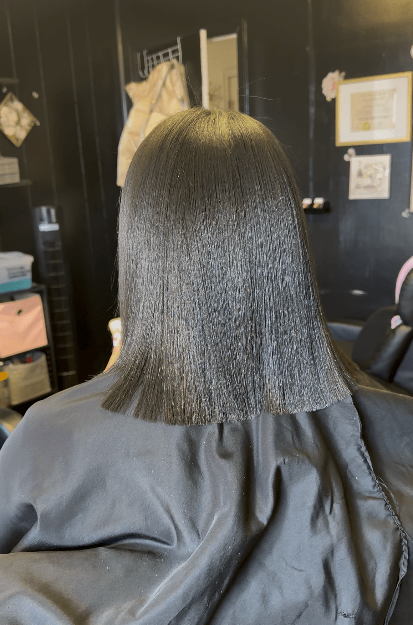 Back of a person with straight, shoulder-length black hair in a salon chair, black cape, dark interior background.