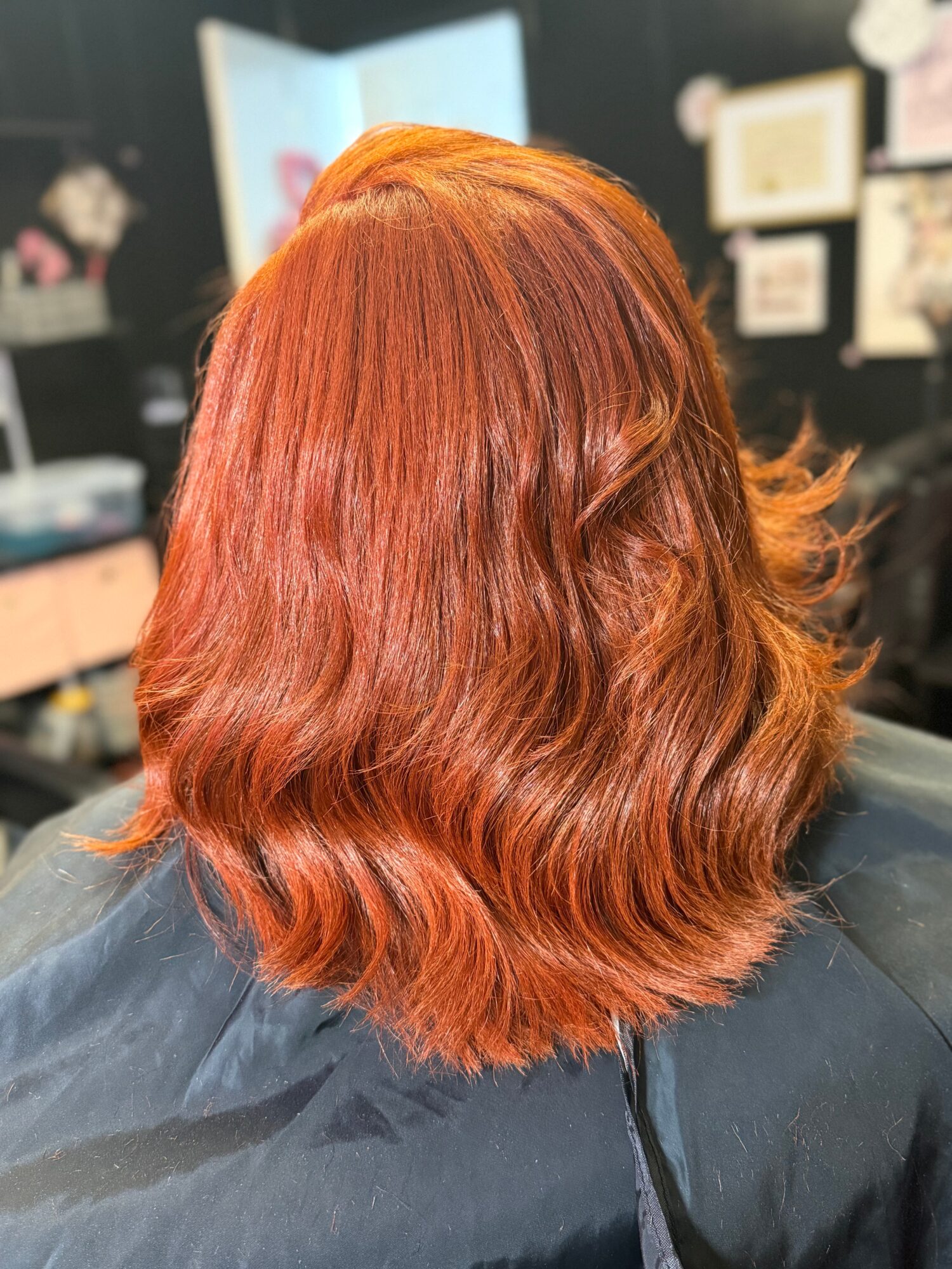 Back of a person's head with wavy, shoulder-length, reddish-orange hair in a salon setting.