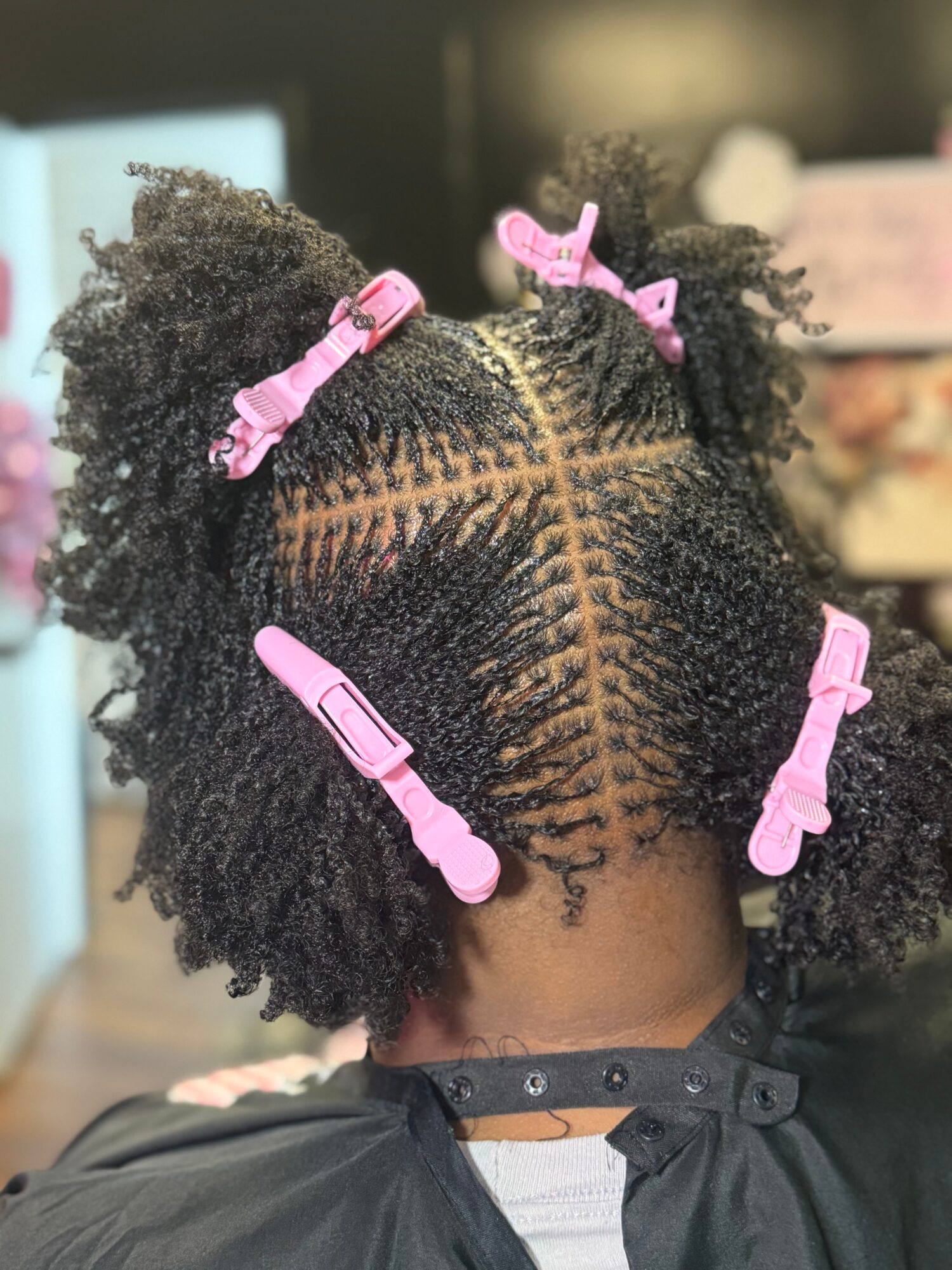 Back of a person's head with natural curly hair styled in two puffs secured with pink clips, showing a parting pattern.