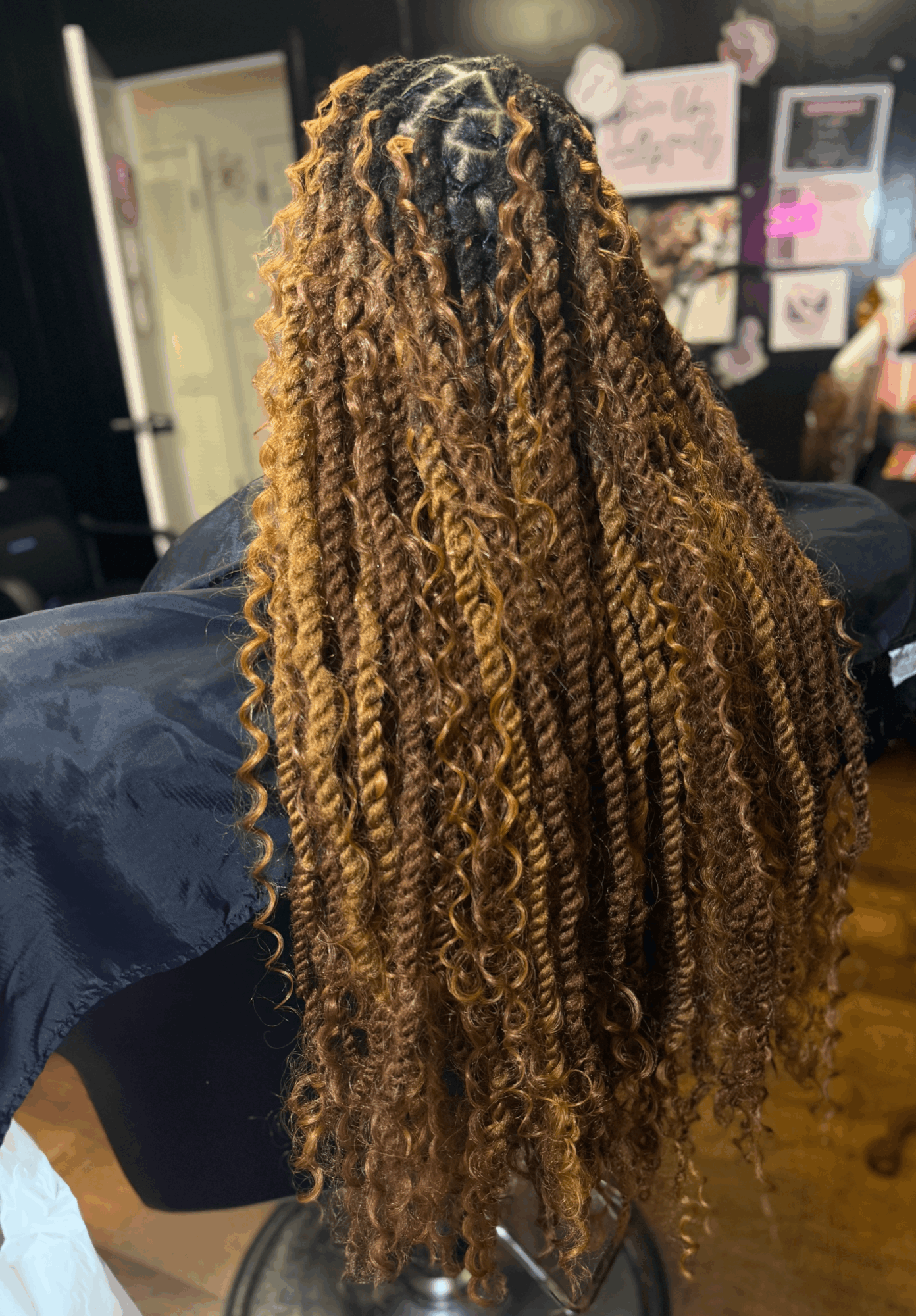 Person with long, curly, two-tone hair in a salon or similar setting, with posters and photos on the wall behind.