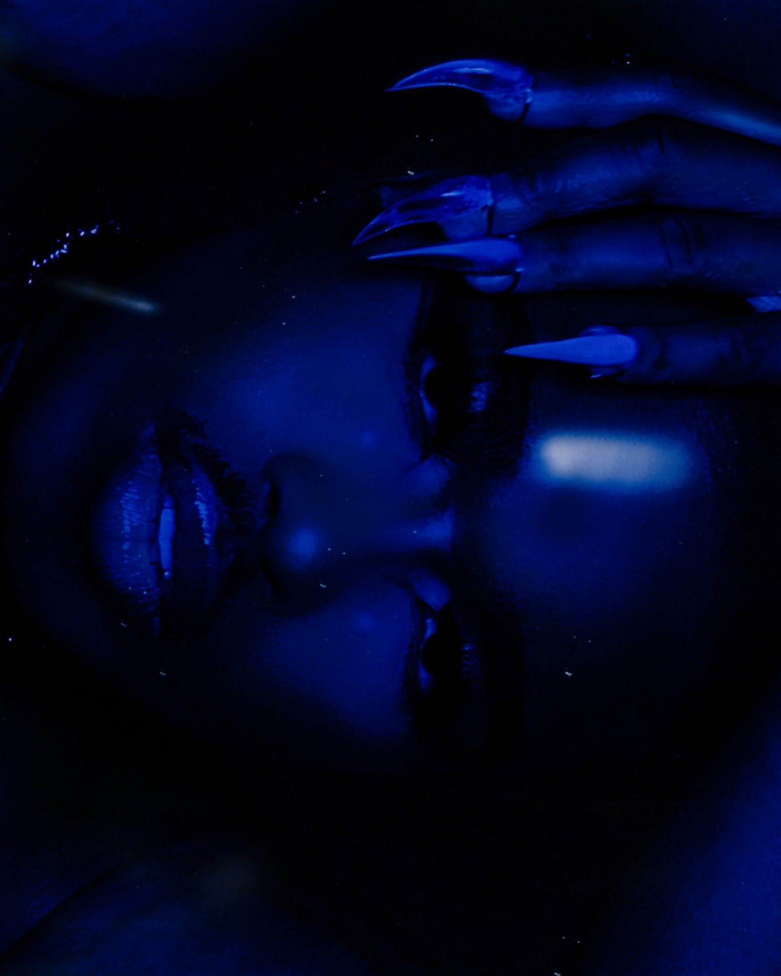 Close-up of a person's face with glowing blue makeup and long nails, touching their lips in a dark setting.