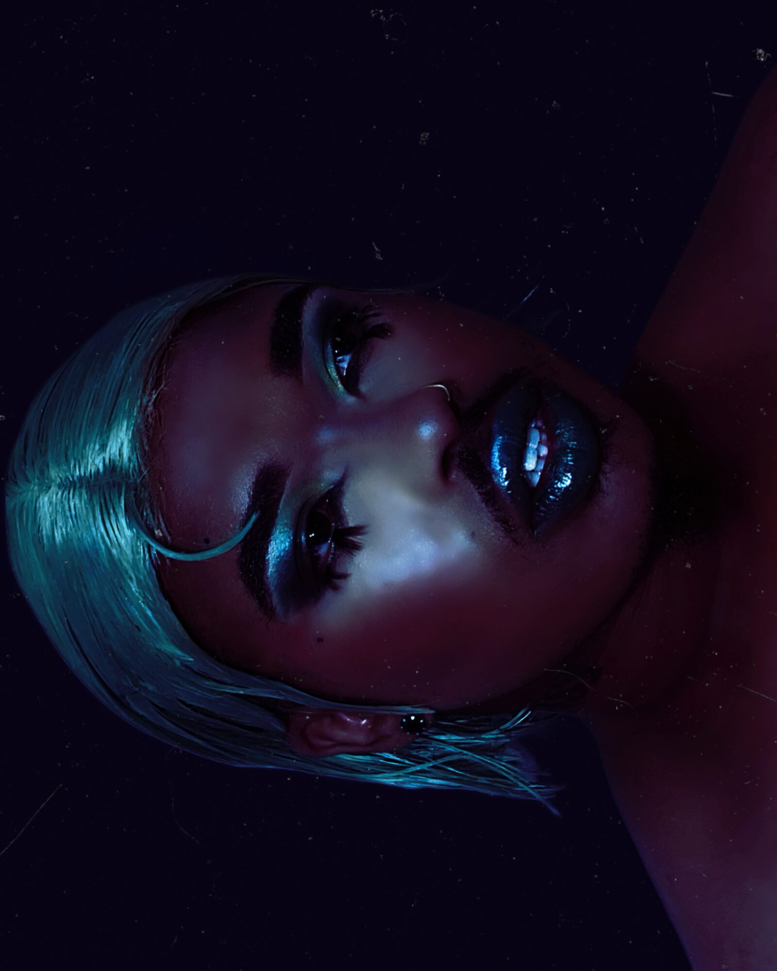 Close-up of a person with metallic makeup and hair, lying in dark surroundings, looking upward with dramatic lighting.